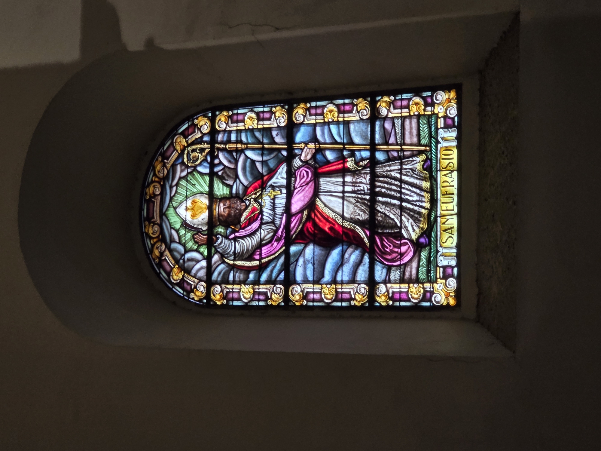 This image shows a stained glass window depicting a religious figure, likely a saint or bishop, wearing a mitre and holding a crosier. The figure is dressed in ornate robes of red, white, and pink, with a golden cross around its neck. The stained glass is set within an arched window frame and is surrounded by decorative borders. Below the figure, text in Latin reads "SAN EUFRASIO," identifying the figure as Saint Eufrasius. The window is illuminated from behind, casting a colorful glow. The surrounding environment appears to be the interior of a building, possibly a church or cathedral, with dark, curved walls and a textured sill beneath the window. The light source is diffused and creates shadows on the wall to the right of the window. This stained glass window is located in the Jaén Cathedral, specifically in the Chapel of Saint Eufrasius.