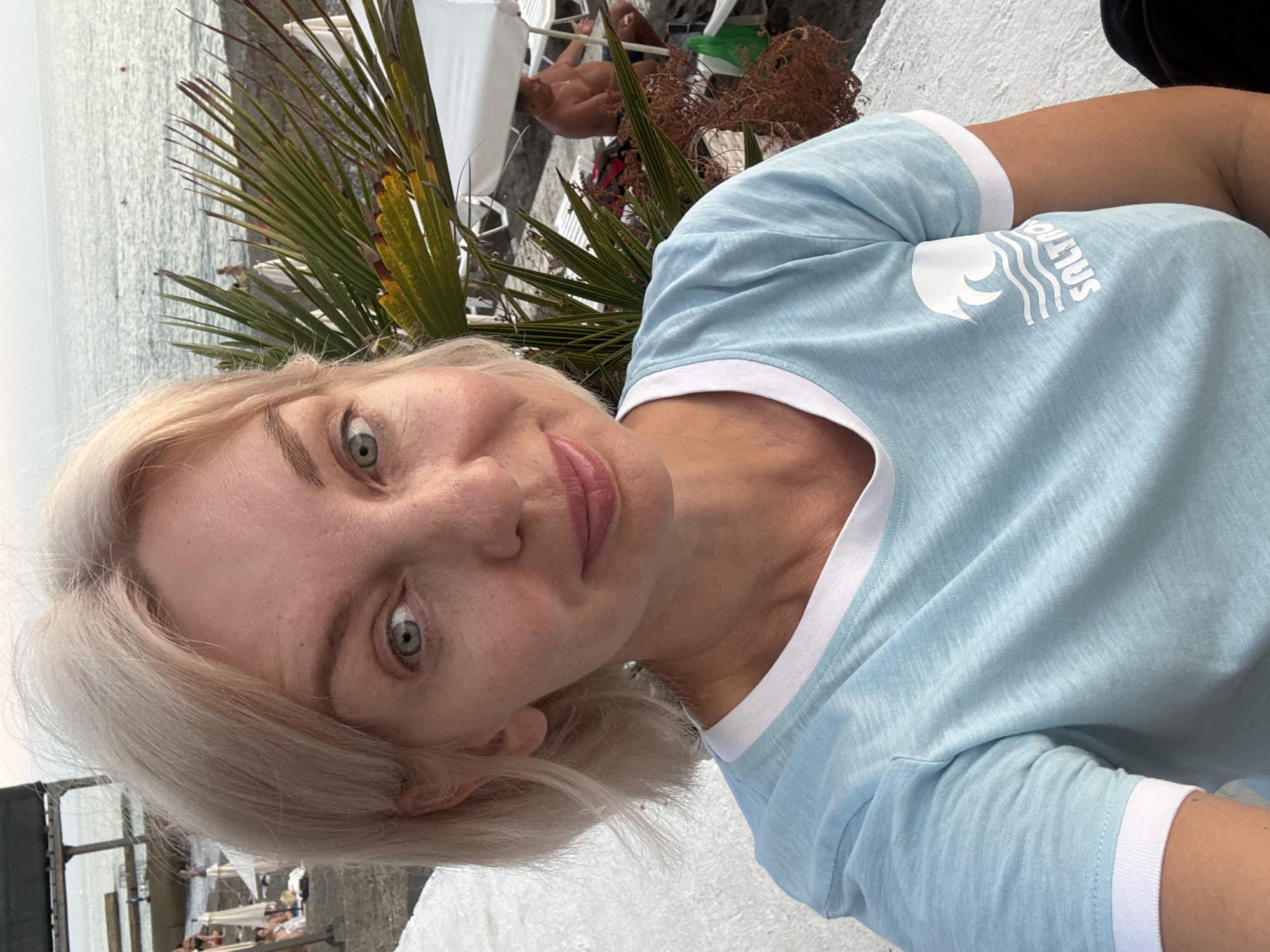 A blonde woman is taking a selfie on a beach in Sochi, Russia. She is wearing a light blue t-shirt with a white collar and cuffs, and a white logo of a wave on the left side of her chest. Her hair is short and styled in loose waves. The background shows the Black Sea, a beach with white lounge chairs and umbrellas, and some palm trees. There are a few other people in the background, some of whom are sitting on the lounge chairs. The lighting suggests it is late afternoon or early evening.