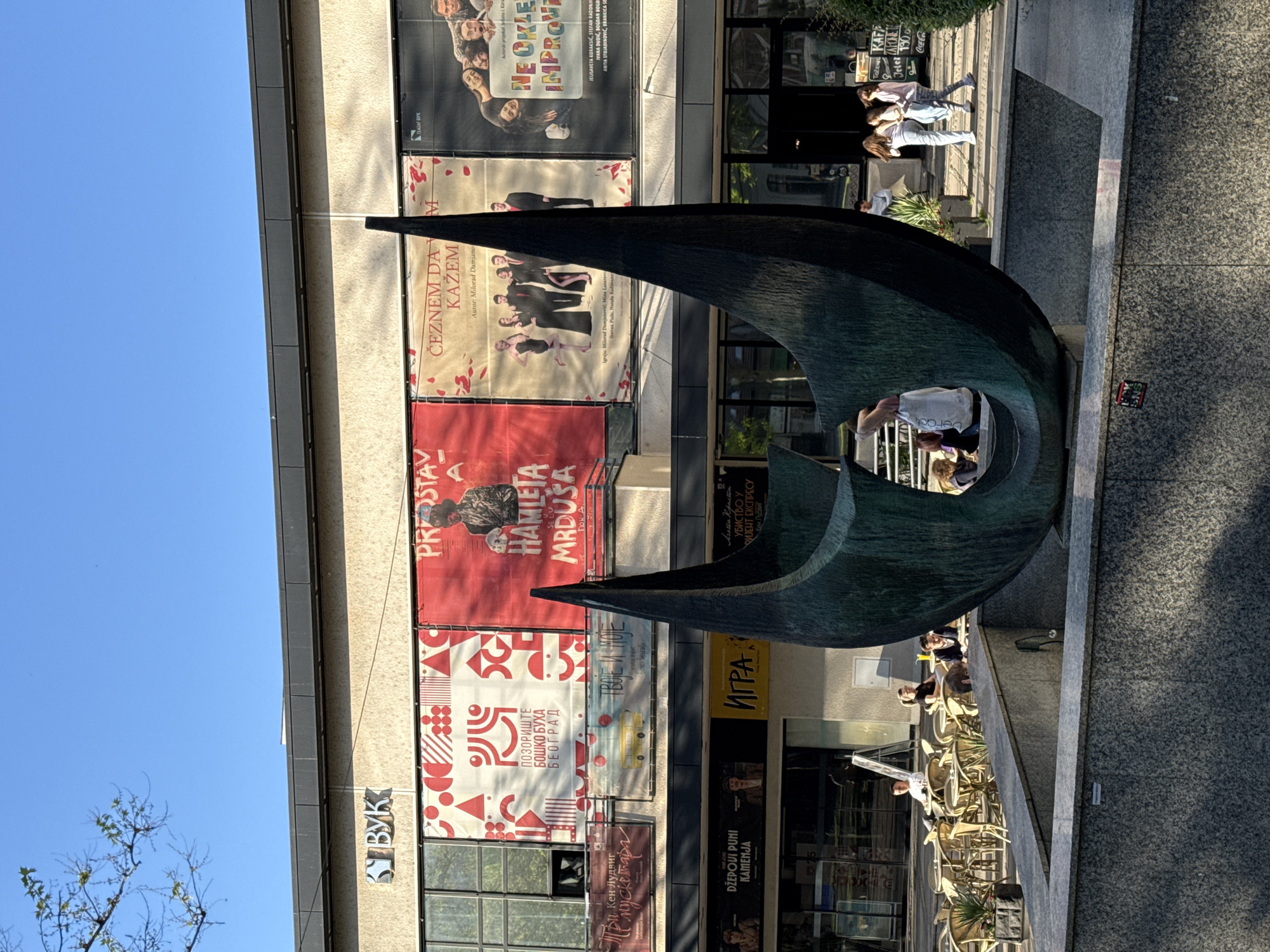 Here is a detailed description of the image:
**Setting and Environment:** The image is taken in an urban environment, likely in the Vracar Urban Municipality, Serbia, based on the Cyrillic script present on signs and posters. The scene is a public square or plaza, with a large, modern sculpture as a central feature. The weather is sunny, casting distinct shadows.
**Architecture:** A tall, modern building forms the backdrop of the scene. The building is covered with large posters for theatrical productions, featuring titles, cast members, and production details in Serbian. Some storefronts are visible at ground level, suggesting commercial or entertainment venues.
**Objects:** The primary object is a large, abstract sculpture made of a dark-colored, possibly metal, material with a textured surface. The sculpture has a curvilinear form, creating an opening or passageway in the center. There are chairs and tables set up near the sculpture, suggesting a cafe or seating area.
**People:** Several people are present in the scene:
* Individuals are walking on the sidewalk adjacent to the building.
* People are seated at the cafe tables.
* A person is walking through the sculpture's opening.
**Actions and Interactions:** People are engaged in various activities, including walking, sitting, and interacting with the sculpture. The sculpture appears to be a focal point for people to gather or pass through.
**Overall Impression:** The image captures a vibrant public space in an urban setting, where art and culture intersect with everyday life. The modern sculpture acts as a landmark and social hub.