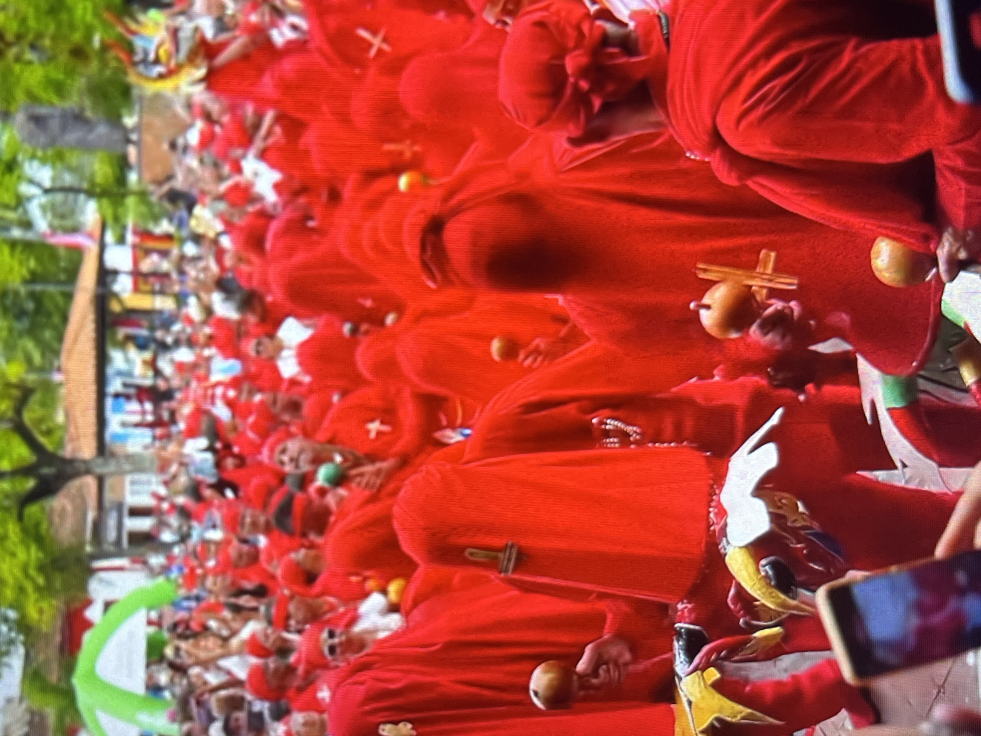 The image depicts a street scene in Tinaquillo, Venezuela, during a procession or festival. A crowd of people, many of whom are wearing bright red hooded robes, are participating. Some individuals are holding maraca-like instruments, while others are holding what appear to be small golden objects. In the foreground, a person is holding up a smartphone to take a picture. In the background, there are buildings with tiled roofs, trees, and a statue. The overall atmosphere suggests a public event with a strong cultural or religious theme, characterized by the prominent use of the color red and traditional attire.