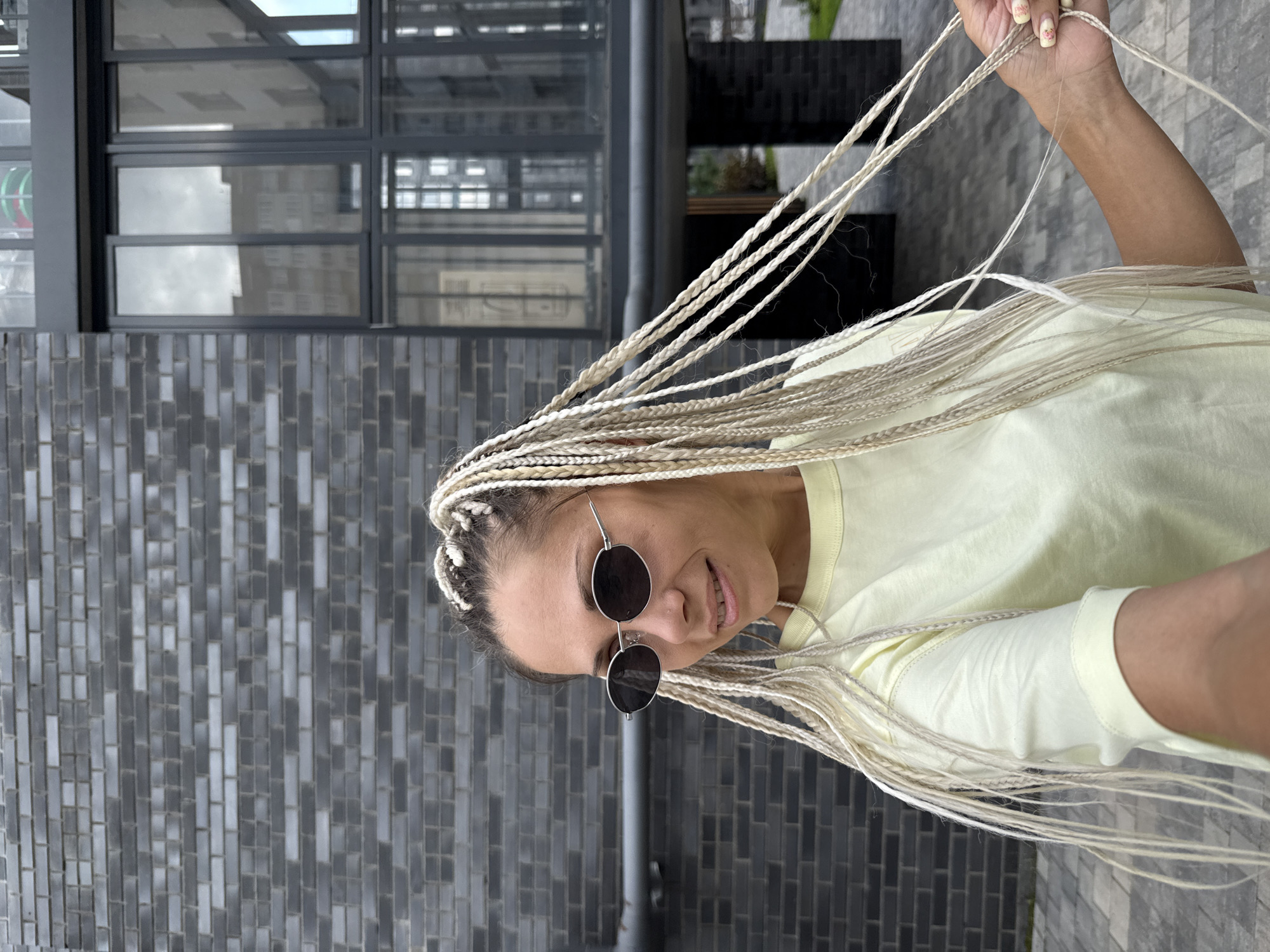 A woman with blonde braided hair and sunglasses is standing in front of a dark brick building. She is wearing a light yellow t-shirt. Her right hand is holding some of her braids. The background shows a modern building with large windows. The scene appears to be outdoors in an urban setting, likely in the city of Rusanovo, Russia.