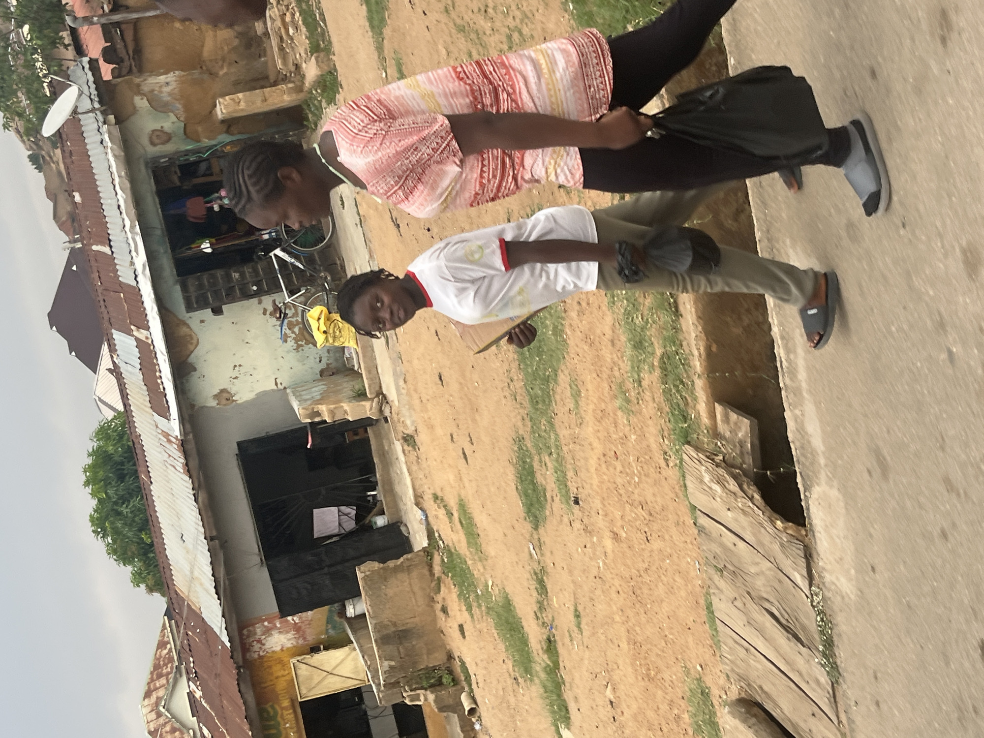Two Black women are walking on a street in Jos, Nigeria. The woman closer to the camera is wearing a white t-shirt with red trim, olive green pants, and grey slides. She is holding a cell phone. The other woman is wearing a pink and white striped top, black pants, and grey slides. She is carrying a black plastic bag. The background includes a building with a rusty corrugated metal roof and a satellite dish. The ground is a mix of dirt and grass.