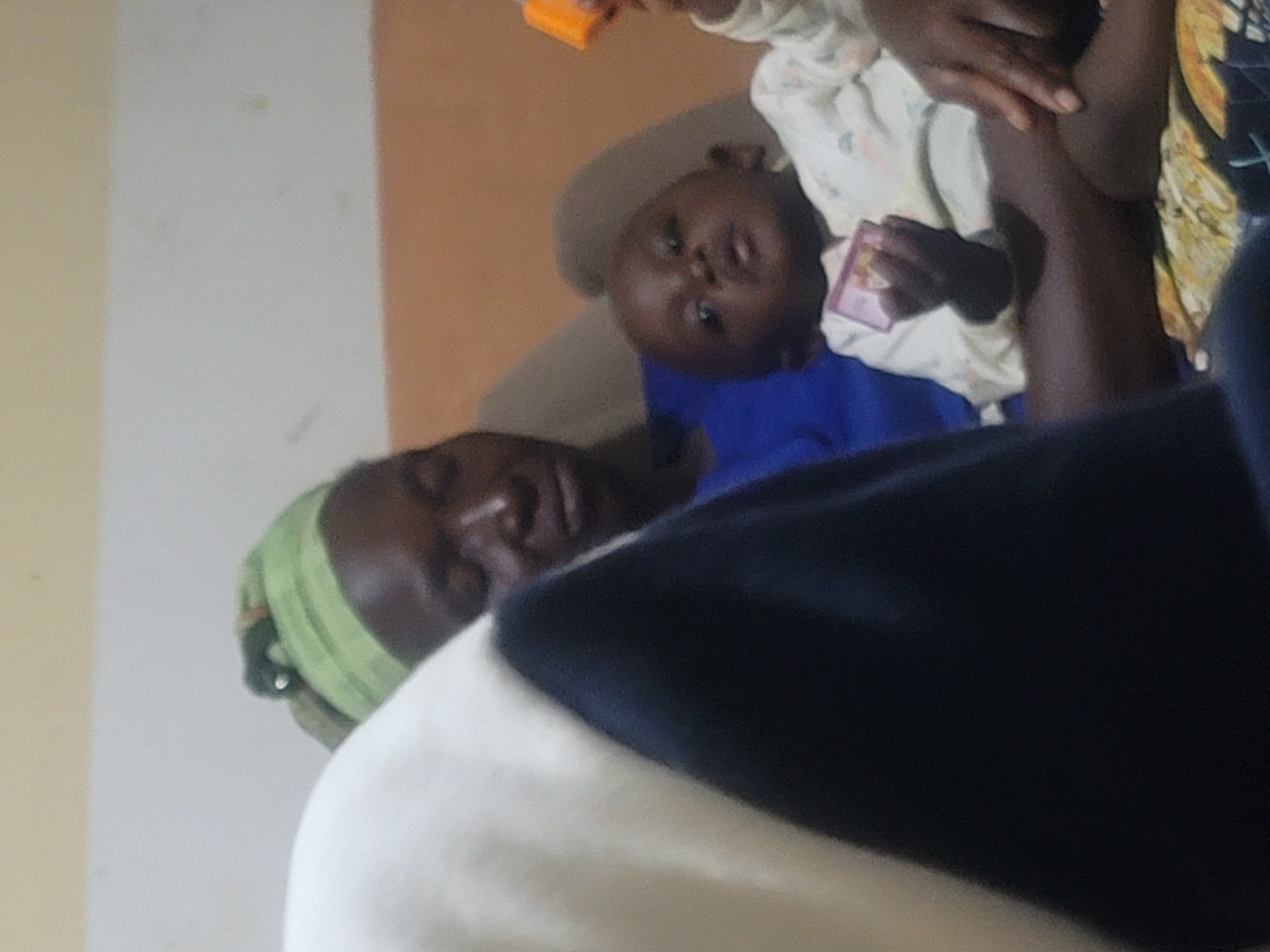 In an indoor setting within the city of Unknown, Unknown, a dark-skinned adult holds a dark-skinned infant. The adult wears a light green headwrap, secured with a small white circular object near the forehead, and a blue garment. Their eyes appear closed or downcast. The infant, wearing a light-colored, long-sleeved top with faint patterns, sits on the adult's lap and looks forward. The infant holds a small orange toy block in their raised right hand and a small pink/purple rectangular object (possibly a card or toy) in their left hand. The adult's right hand rests on the infant's left thigh. The pair is seated on a beige-upholstered surface, covered by a patterned fabric in yellow, brown, and blue beneath the infant. The background features a wall painted with horizontal stripes: a light yellow-beige top, a light gray middle with minor smudges, and a reddish-brown lower section. A large, blurry white and dark blue object partially obstructs the left foreground.