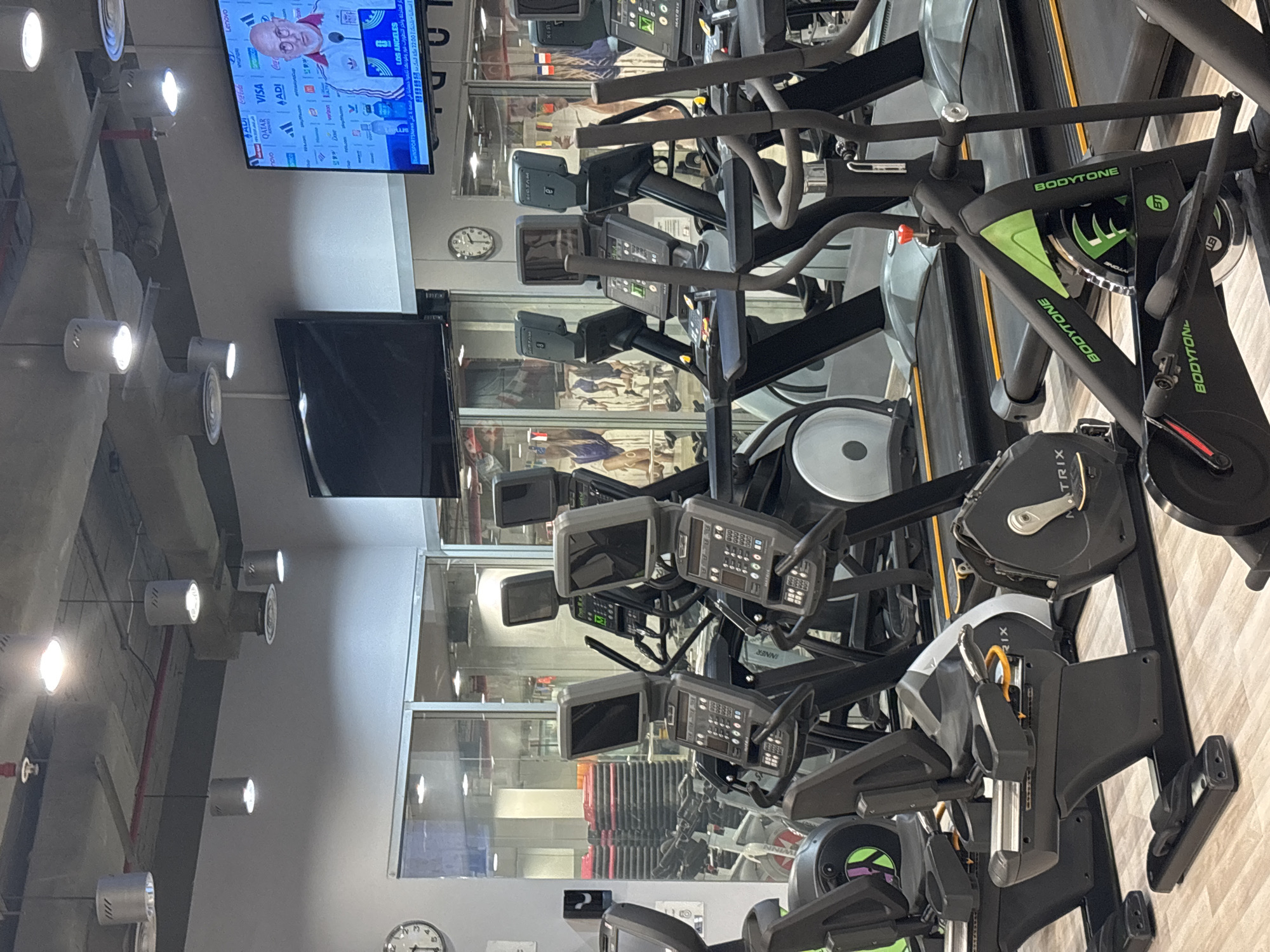 This image depicts the interior of a modern gym, likely located in Amman, Jordan, characterized by rows of exercise equipment, including treadmills, elliptical machines, and stationary bikes. The environment is brightly lit with recessed ceiling lights and illuminated by the screens of various entertainment systems and televisions mounted on the walls. One prominent television displays a broadcast with Arabic text and images of sports-related content, suggesting a connection to a sporting event. The gym appears to be clean and well-maintained, with reflective glass partitions allowing views into other sections of the facility, revealing more exercise machines and what appear to be stacked aerobic steps. The overall impression is one of a functional and technologically equipped fitness center, ready for use.