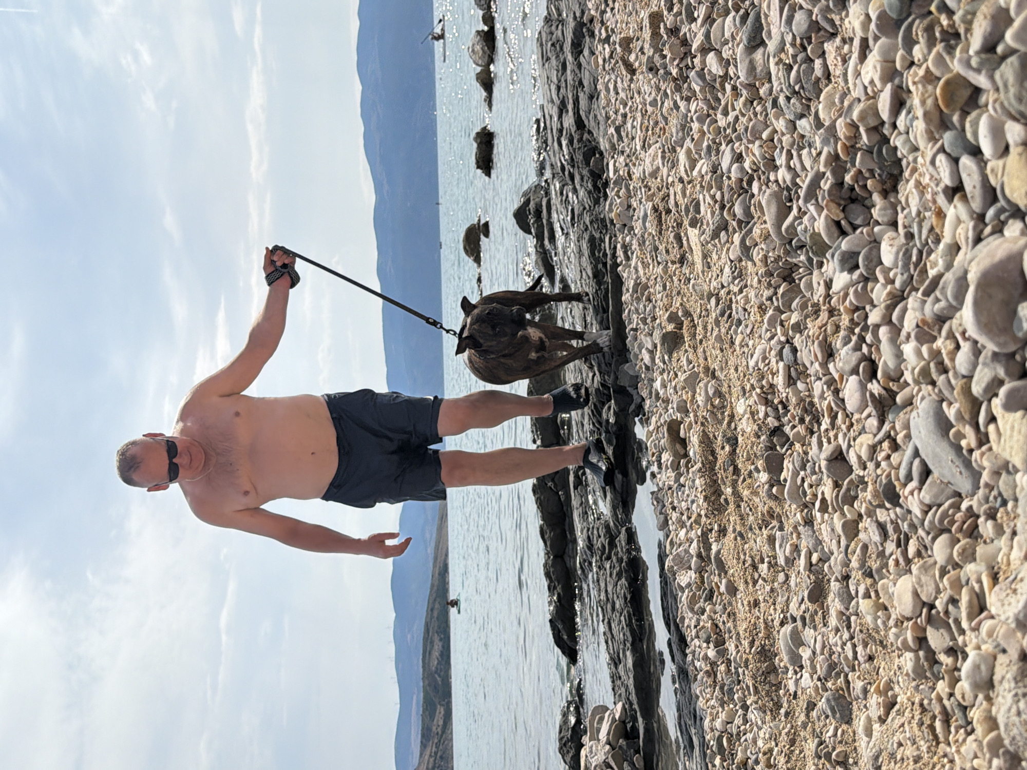 An shirtless man wearing dark swim shorts and sunglasses walks a brindle-colored pit bull terrier mix on a leash along a rocky shoreline with the Adriatic Sea in the background. The man is bald and has a chain necklace. The dog is walking in front of him, looking to the left. In the distance, several other people are visible on the water, including one on a paddleboard and two in what appear to be kayaks. The sky is partly cloudy with a few cumulus clouds. The background features a mountainous, sparsely vegetated coastline. The terrain in the foreground is comprised of small to medium-sized pebbles and sand. The location is Općina Baška, Croatia.