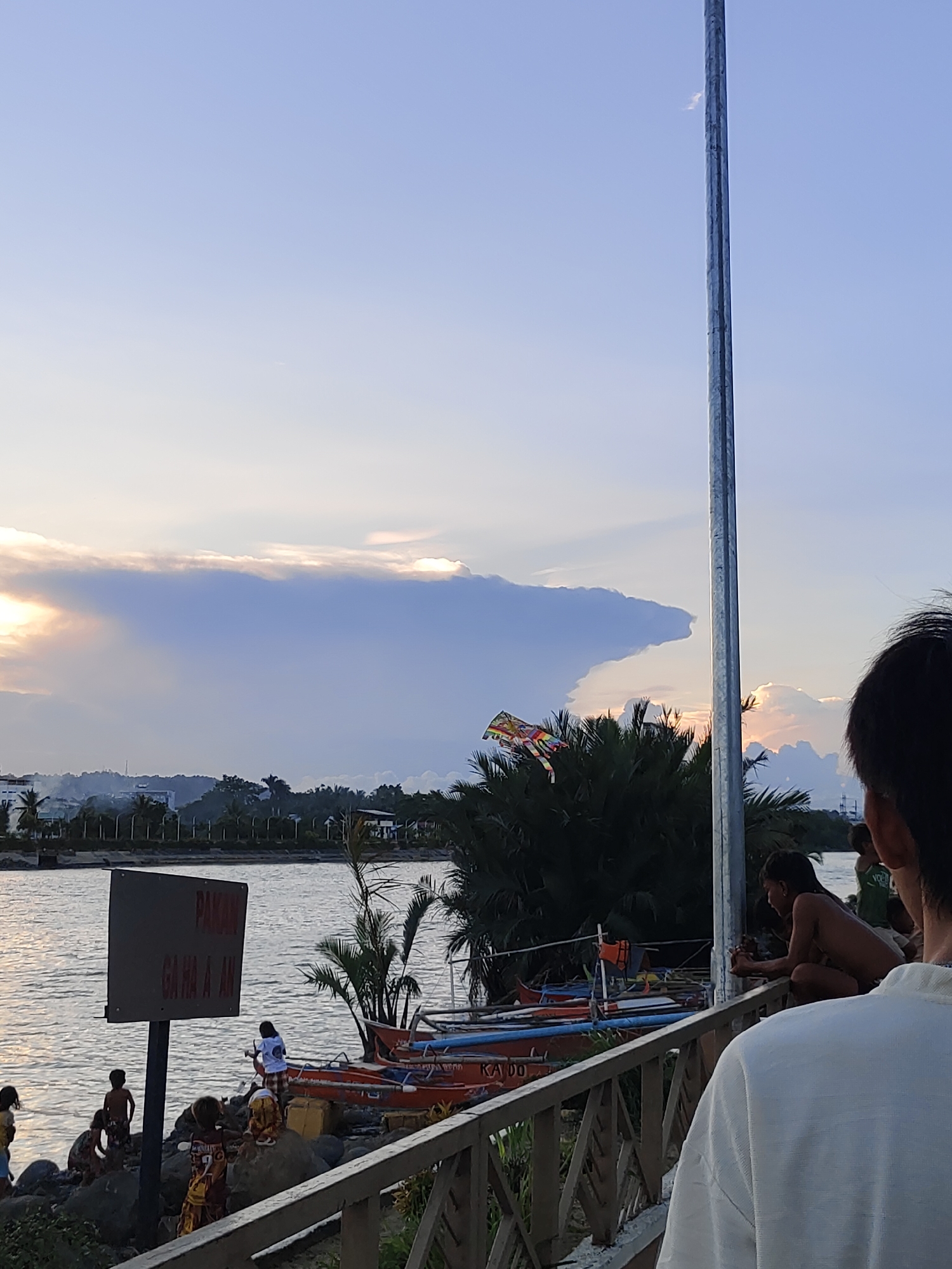 The image depicts a sunset scene at a waterfront in Davao City, Philippines. The sky is a gradient of blue and orange, with large, dramatic clouds illuminated by the setting sun. In the foreground, a wooden railing separates the viewer from the scene. On the left side, a group of children are gathered near the water's edge, some playing on rocks. A sign with red text is partially visible. Several boats are moored near the shore. In the middle ground, palm trees and other foliage are visible, with a kite flying in the distance. The background shows a cityscape with buildings and hills. The overall impression is one of a tranquil evening by the sea.