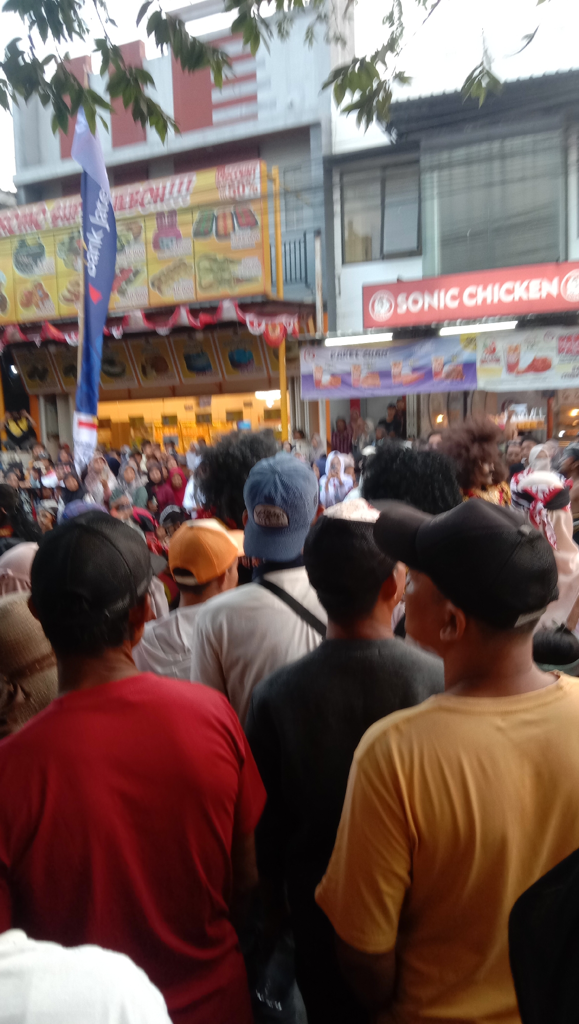 The image shows a bustling outdoor scene in Jatisawit, Indonesia, likely a street or market area. In the foreground, several individuals are seen from behind, wearing casual clothing like t-shirts and hats. The focus is on their backs and heads as they appear to be part of a crowd.
Behind them, a vibrant yellow banner advertising various food items is prominently displayed, with text and images indicating a variety of culinary offerings. A blue flag, possibly associated with a business or event, stands tall next to the banner. Further in the background, a red and white striped building is visible, suggesting an urban or commercial setting. Another sign, with the text "SONIC CHICKEN" in red on a white background, indicates a fast-food establishment.
The crowd appears to be diverse, with some individuals wearing headscarves, common in Indonesia. The atmosphere seems lively and possibly a public gathering or event, given the density of people and the commercial displays. The lighting suggests it is daytime, though the sky is overcast or the image is taken during dusk. The overall impression is of a community event or a busy commercial district in Jatisawit.