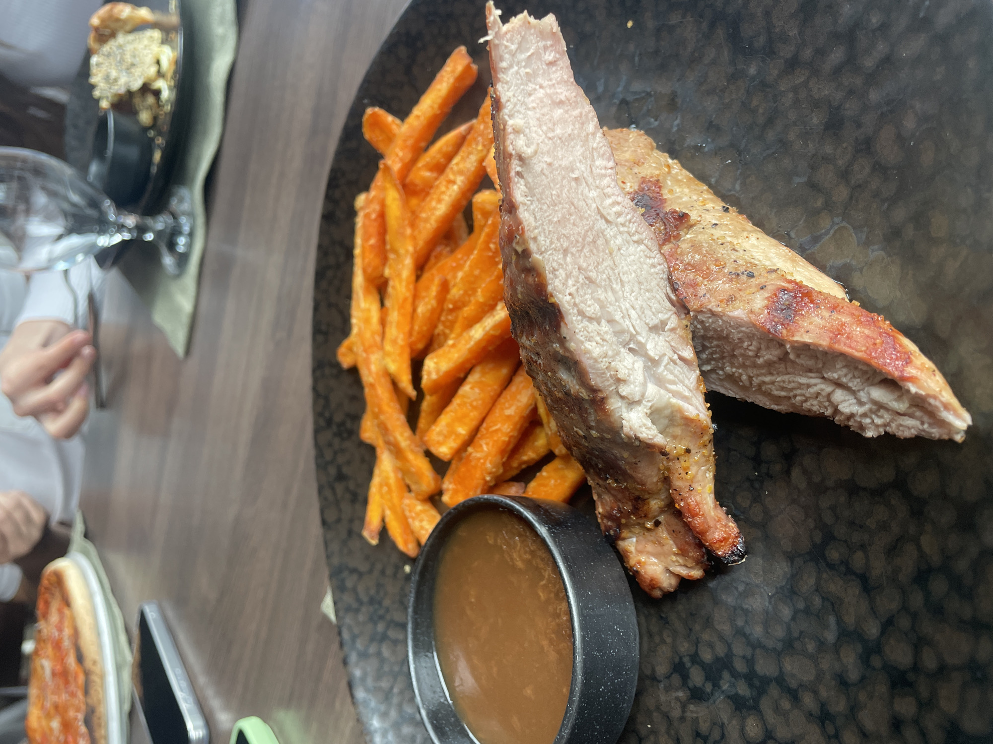 The image shows a black plate containing cooked meat, sweet potato fries, and a small black bowl of brown sauce. The meat is sliced, revealing a cooked interior. The sweet potato fries are arranged to the left of the meat. The black plate has a textured surface. In the background, partially visible, are other dishes, including what appears to be a pizza and a taller dish possibly containing cauliflower gratin. There are also a drinking glass and a person's hand visible in the background. The table surface is wood-grained. The location is Pleven, Bulgaria, but the image provides no information to confirm this.
