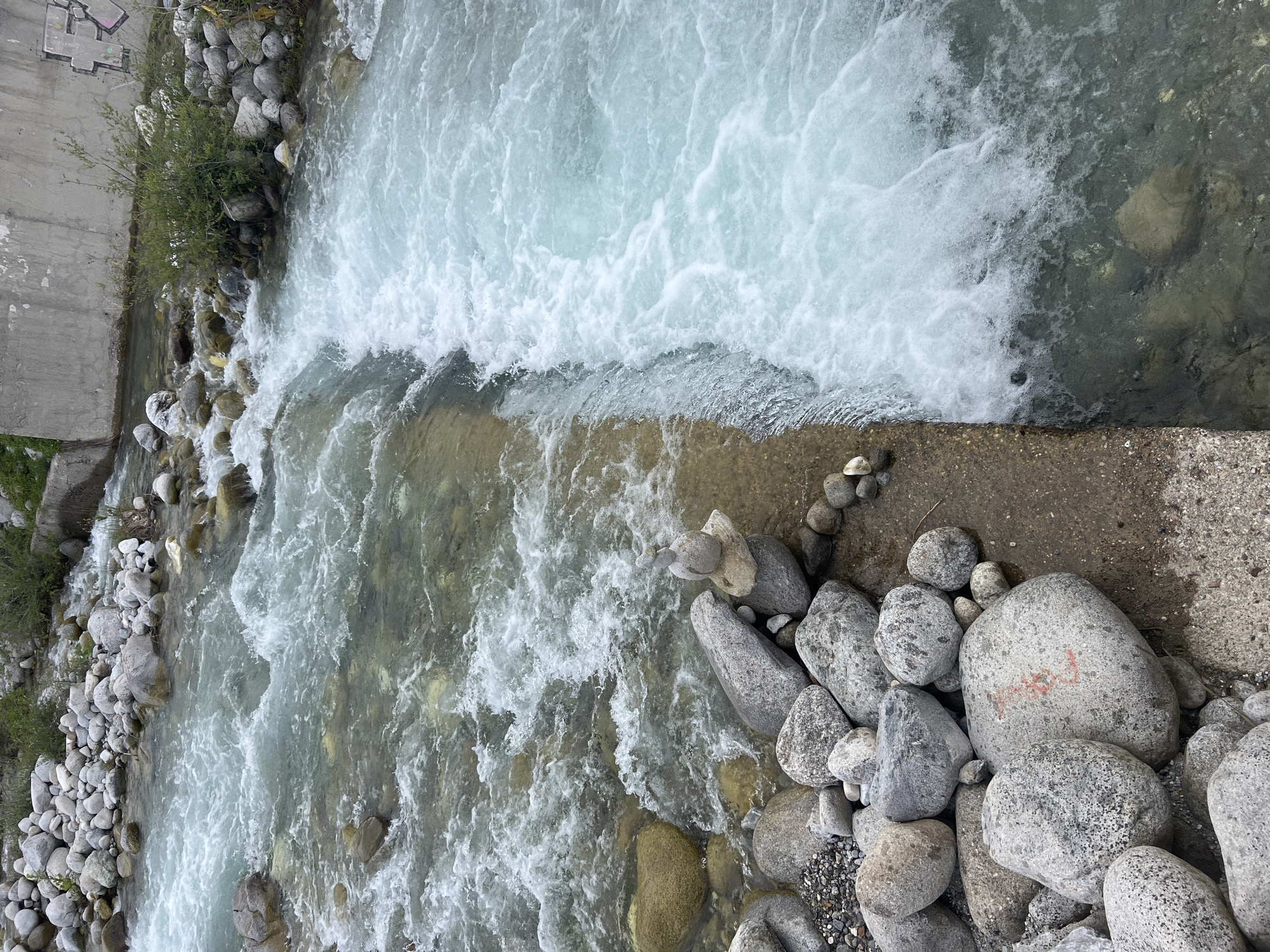 The image captures a natural scene with a river flowing through what appears to be a mountainous or wild environment. The water is clear and turbulent, with white foam indicating rapids or a waterfall. The riverbanks are lined with numerous rocks of varying sizes, many of them smooth and rounded by the water's flow. Some vegetation, consisting of small green bushes and grass, grows along the banks.
In the foreground, on the left side of the image, a collection of rocks forms a distinct arrangement. One prominent large grey rock has red markings that appear to be numbers or symbols. A stack of smaller stones has been carefully balanced on top of another rock, creating a small cairn.
The background shows a concrete wall or structure on the right side, suggesting the presence of man-made infrastructure near the natural landscape. Graffiti can be seen on this wall, adding a contrasting element to the natural setting. The overall impression is of a wild river, possibly within or near a natural park or undeveloped area, with elements of human presence in the form of the concrete wall and graffiti. There are no people visible in the image.