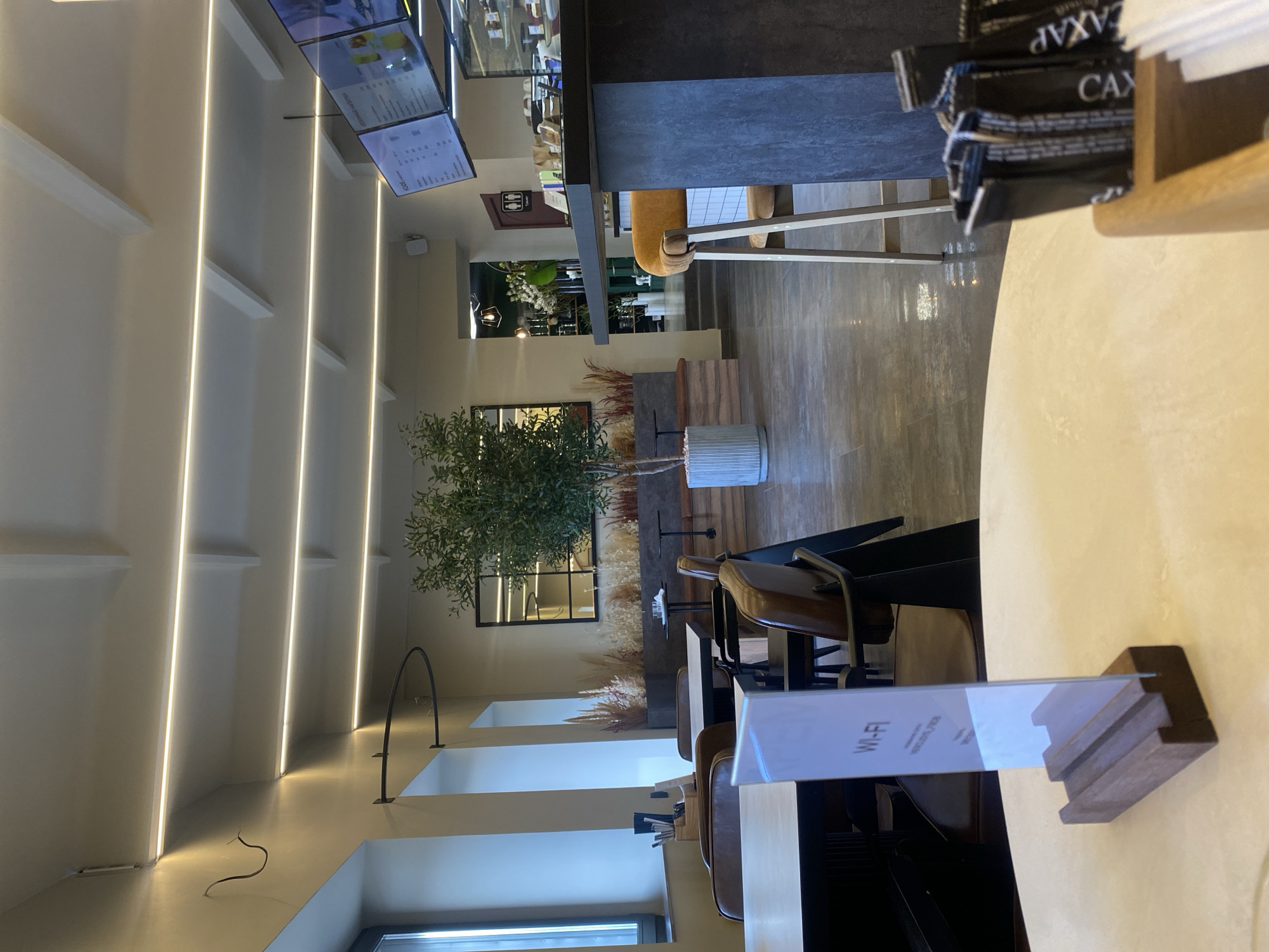 The image depicts the interior of a modern cafe or restaurant, likely located in Votkinsk, Russia, given the context of the request. The scene is well-lit with overhead linear LED lighting strip illuminating the ceiling and upper walls.
In the foreground, a light-colored round table is visible, with a transparent "Wi-Fi" sign in a wooden stand placed on it. To the right of the table, a wooden box contains several packets of sugar, one of which is partially visible with the word "SAXAP" (sugar in Russian) printed on it.
Further into the cafe, there are several seating areas. A dark-colored bar or counter dominates the right side of the image, with a tall orange stool with wooden legs pulled up to it. Behind the counter, shelves are stocked with various items, and a display case likely holds food items. Digital menu boards are mounted on the wall behind the counter, showing pricing and descriptions in Russian.
In the middle ground, several tables with dark chairs are arranged. One table is visible on the left, with a wooden organizer holding pens or utensils. Another seating area features brown leather chairs around tables, with a large potted plant adding a touch of greenery to the space. Beyond this, a long bench seating area is visible against a wall, with small tables set up.
The background reveals more of the cafe's interior, including a doorway leading to another section, possibly a kitchen or restrooms, indicated by a sign with two figures. The overall ambiance is contemporary and clean, with a mix of natural materials like wood and concrete, and polished floors. No people are clearly visible in the scene, suggesting the photo was taken during a quiet period. The decor includes elements like dried grass arrangements and a large mirror reflecting the ceiling lights.