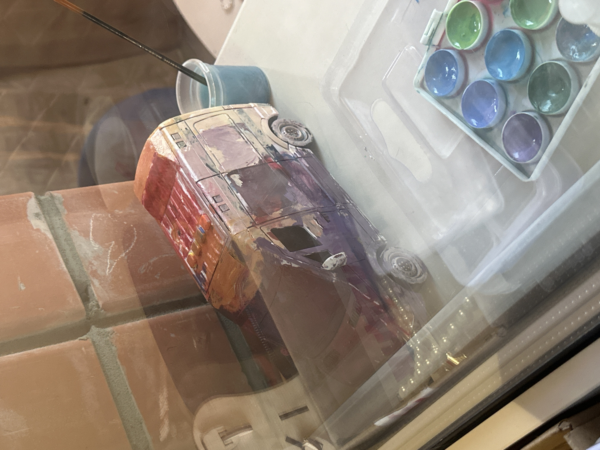 The image shows a detailed, hand-painted model of a van. The van is predominantly purple and red with streaks of other colors, suggesting an artistic or weathered effect. It is positioned on a light-colored surface, possibly a table or shelf.
To the right of the van, a clear plastic cup filled with blue liquid is visible, with a paintbrush resting inside. In the foreground, a white plastic tray contains several small, round wells filled with various colors of paint, including shades of blue, purple, and green.
In the background, a brick wall with a textured surface and green mortar is apparent. The lighting suggests an indoor environment, possibly a workshop or art studio.
The provided context states the image is from Saransk, Russia. However, there are no specific visual cues within the image that directly indicate its location within the city of Saransk. The scene depicts a close-up view of crafting supplies and a model, rather than an identifiable cityscape or landmark.