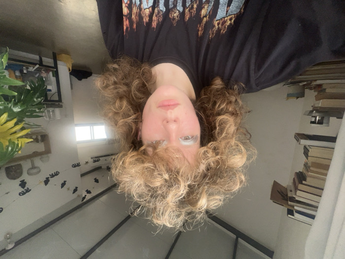 A young adult female is depicted centrally within an indoor setting in Tel-Aviv, Israel. She has voluminous, light brown curly hair, styled with significant upward lift. Her eyelids feature light blue eyeshadow. She maintains a neutral facial expression while looking directly forward. She wears a black short-sleeved t-shirt with a multi-colored graphic design across the chest.
The room features white walls and a light-colored floor, possibly concrete. The ceiling shows dark, exposed structural beams. To the subject's left, a tall stack of approximately 20-30 books rests on a shelf, partially obscured by a light-colored curtain. To the subject's right, a multi-pane window admits bright daylight, below which a dark object rests on the floor. An air conditioning unit is mounted above the window. A decorative string of black cutout shapes is affixed to the wall above the air conditioner. Further right, a multi-tiered wooden shelf displays various objects, including a white mushroom-shaped item. A dark table holds a clear vessel and other items. A large, green plant with some yellow foliage partially obscures the rightmost background.