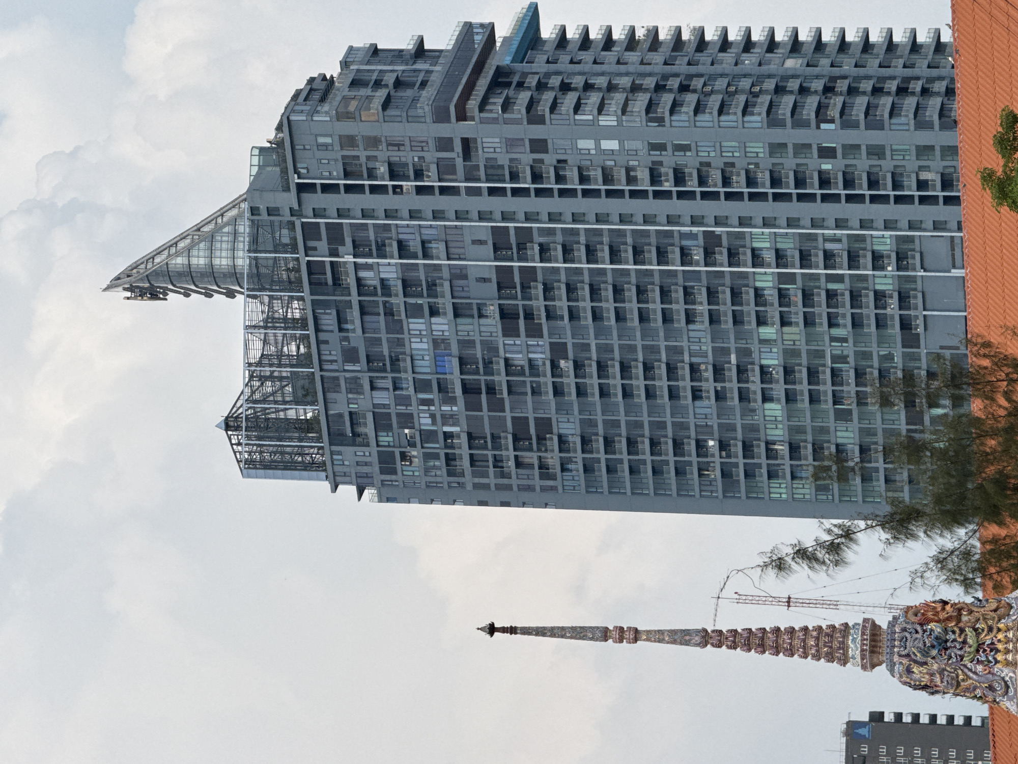 The image shows a tall, modern, gray building located in Bangkok, Thailand. The building has many windows and a unique design with a cantilevered structure at the top. In the foreground, there is an ornate Thai-style spire, typical of temples in the area. The sky is cloudy. There is no evidence of people visible.