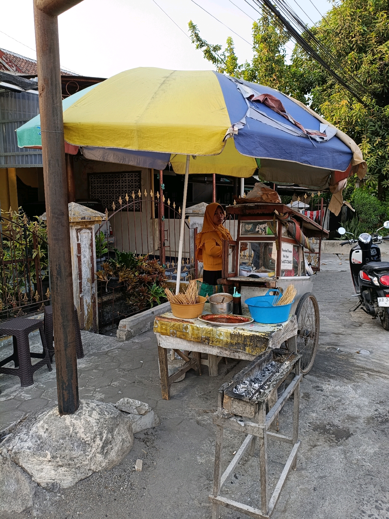 In Purwomartani, Indonesia, a street vendor operates a food cart under a large, worn umbrella. The vendor, a woman wearing a yellow hijab and orange top, stands beside the cart. The cart itself is a mobile unit with a display case and various food items, including skewers in a blue bucket, bowls of ingredients, and a tray with a sauce. A charcoal grill sits in front of the cart. A motorcycle is parked to the right of the vendor. The setting appears to be a sidewalk or street with a house and greenery in the background. The overall scene depicts a typical roadside food stall.