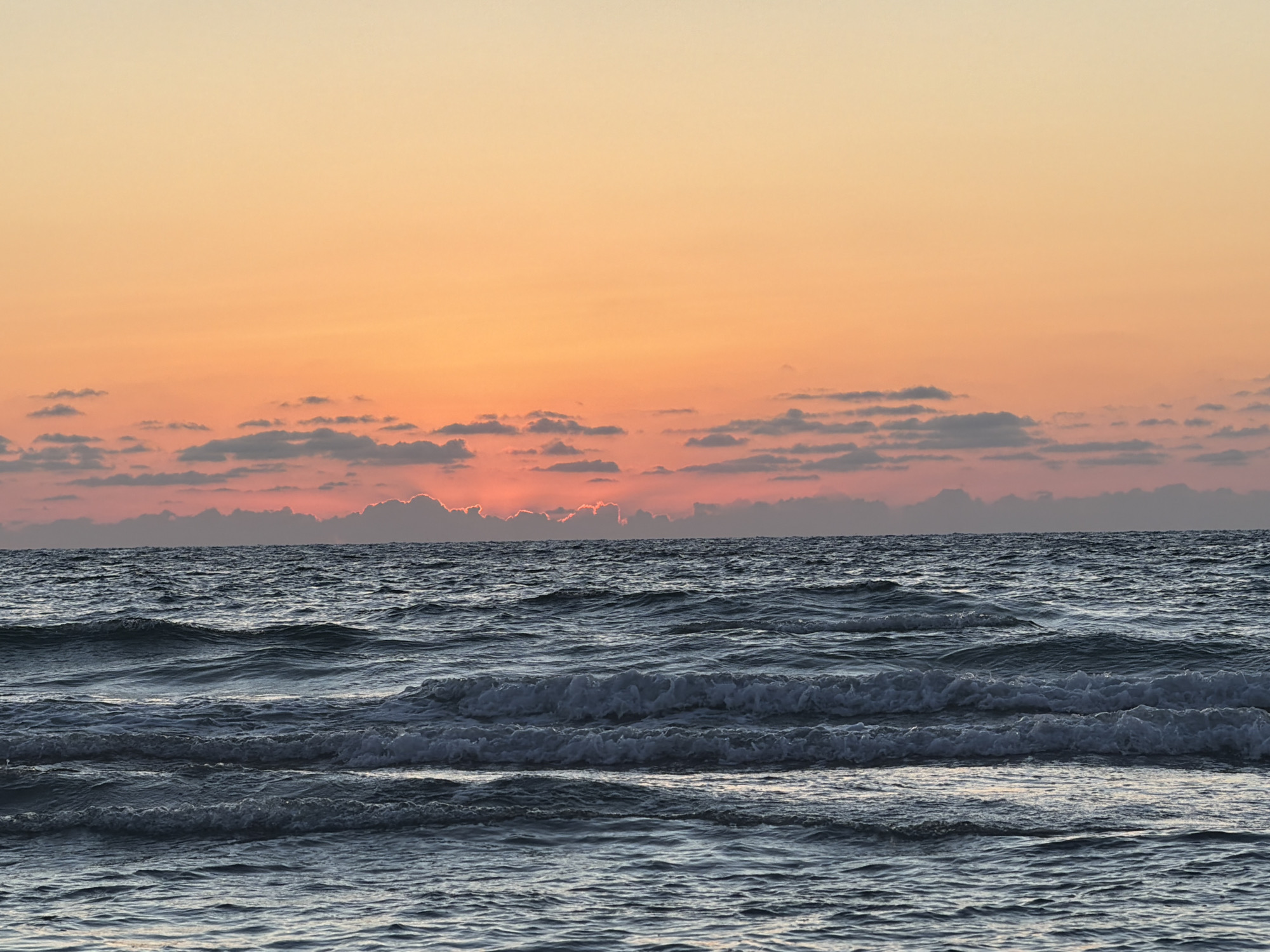 The image depicts a seascape at sunset. The sky transitions from a light orange at the top to a darker pinkish-orange near the horizon, where a line of clouds obscures part of the setting sun. Below the horizon, the sea is visible, with waves rolling toward the viewer. The water is a dark blue-gray color. The image was likely taken in Tel-Aviv, Israel, along the coast, as the city is known for its beaches and sunsets over the Mediterranean Sea. There are no people visible in the image.