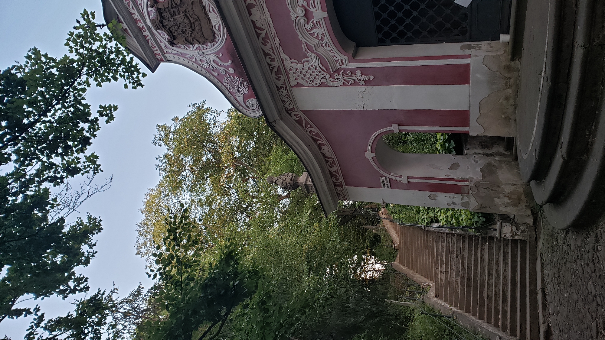 The image shows a close-up of a pink and white baroque-style building with ornate decorations. A steep, stone staircase with a metal railing is visible to the left, leading upwards into a dense green forest. The building appears to be an entrance or gateway to a historical site, possibly a castle or religious complex. The overall atmosphere is serene and slightly melancholic, with the weathered facade of the building and the overhanging trees suggesting age and a sense of quiet seclusion. This building is part of the Calvary in Prešov, Slovakia, a prominent historical and pilgrimage site located on a hill overlooking the city.