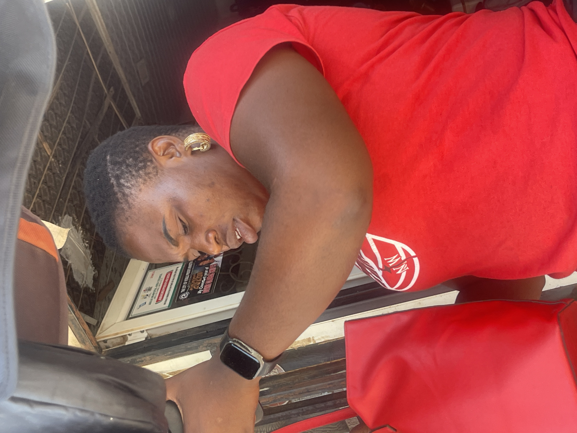 A woman in a red t-shirt with a white logo is holding a red bag. She is wearing gold hoop earrings and a smartwatch on her left wrist. The background shows a shop with signs in what appears to be English, suggesting an urban environment, possibly Abuja, Nigeria, given the context. The lighting suggests it is daytime.