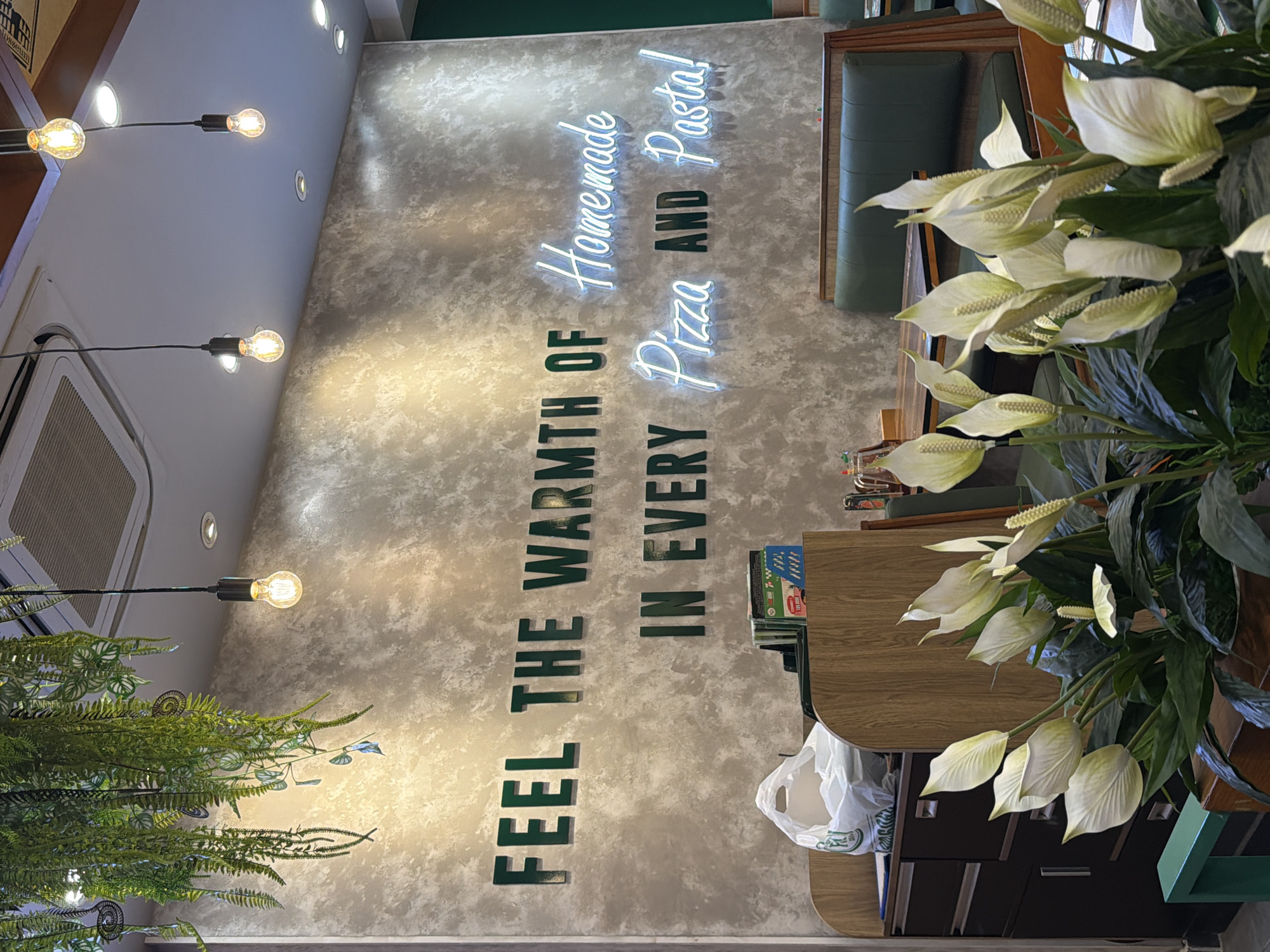 The image depicts an interior space, likely a restaurant, with a decorative wall featuring the text "FEEL THE WARMTH OF" in dark green vertical letters, followed by "IN EVERY" also in dark green vertical letters. Above this, there is a neon sign that reads "Homemade Pizza and Pasta!" The wall itself has a textured, mottled gray appearance. To the left, a hanging light fixture with exposed bulbs is visible, as well as a fern-like plant. To the right, a table or counter is partially visible, with a stack of books and a white plastic bag. A large Peace Lily plant with white flowers occupies the right foreground, partially obscuring the view. The presence of the text and design suggests the image was taken in an eatery; however, information on the city of Đà Nẵng, Vietnam is not visible, and it cannot be determined.