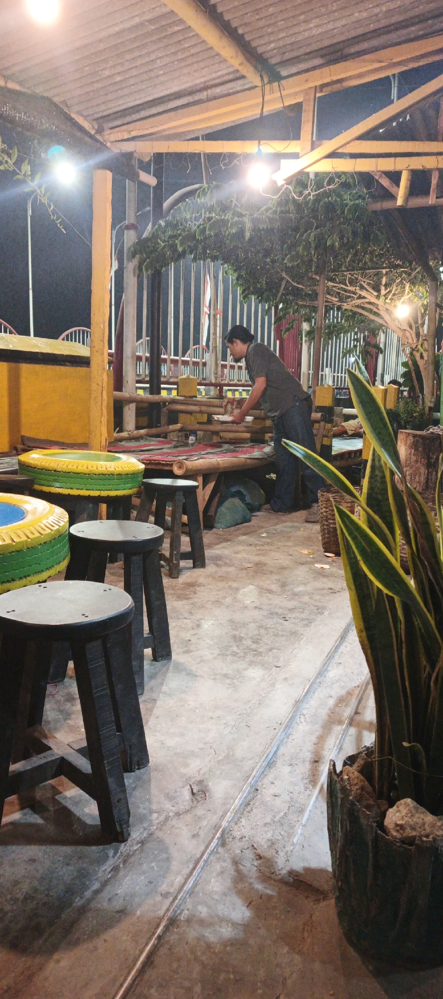 A man is standing at a bamboo table in an outdoor seating area. He is wearing a dark gray t-shirt and blue jeans. The table is covered with a red and black striped mat. There are several wooden stools around the table, some made from tires painted yellow and green. The seating area appears to be part of a restaurant or cafe in Kasiyan, Indonesia, with a roof made of corrugated metal supported by wooden beams and yellow pillars. In the background, there is a fence and some trees. A snake plant in a concrete pot is in the foreground on the right side of the image. The ground is concrete. The scene is lit by artificial lights, suggesting it is nighttime or dusk.