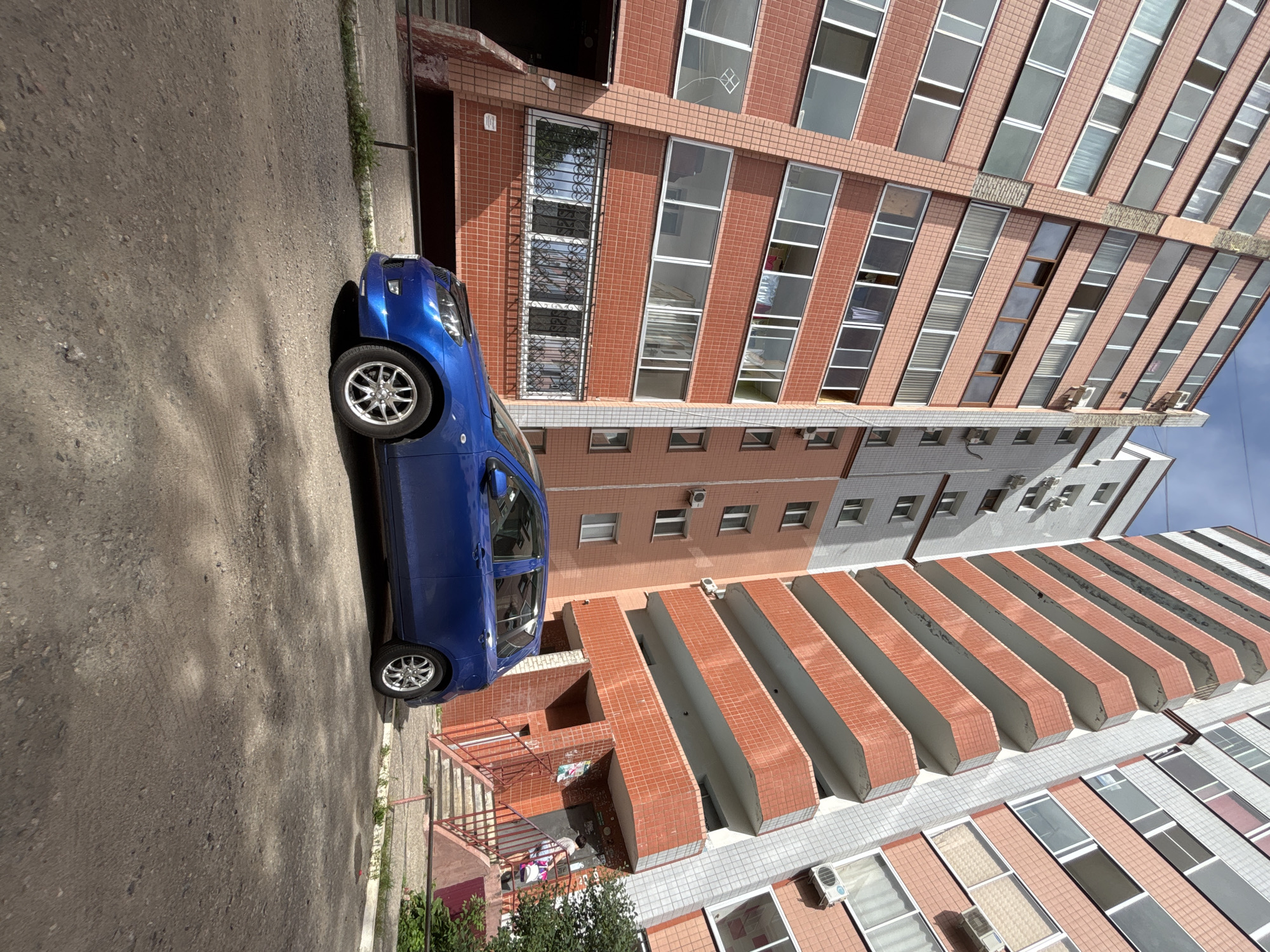 A blue hatchback is parked on a gravel surface in front of a tall apartment building. The building has a facade of small, reddish-brown tiles and white and gray sections. Balconies with reddish-brown tile detailing are visible on the right side of the building. A second, taller apartment building with a lighter gray and white facade is visible to the left of the primary building. A set of stairs leads up to an entrance of the building on the right. A person is ascending the stairs with a white bag. The sky above is partly cloudy. The scene is likely located in Blagoveshchensk, Russia, based on the architectural style and common urban setting.