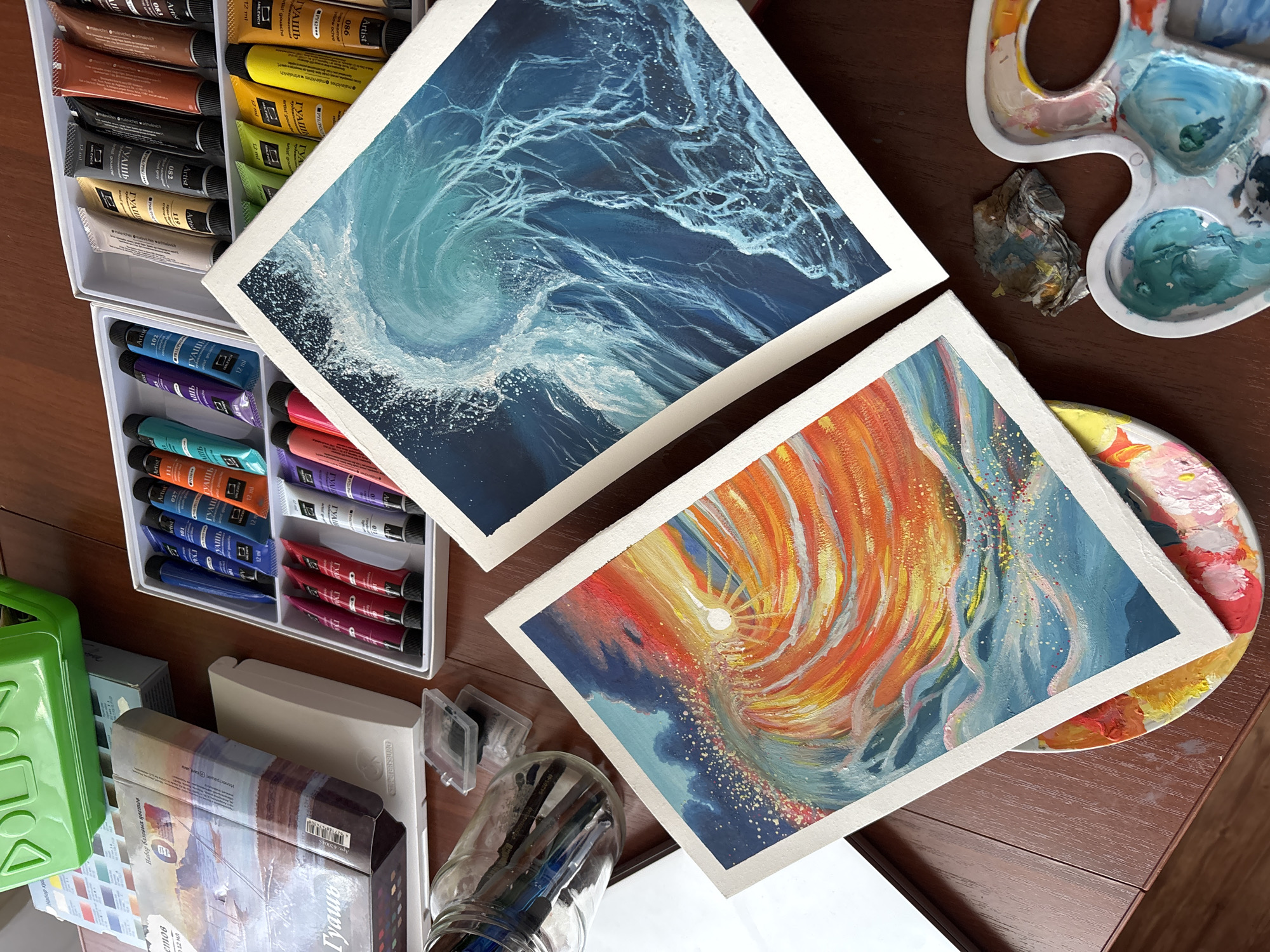 The image is an overhead shot of an artist's workspace, likely in a home setting, as indicated by the wooden table surface. Two completed paintings are centrally positioned. The upper painting features a swirling, dynamic blue and white depiction of an ocean wave. The lower painting showcases an abstract swirl of orange and yellow hues, with a sun or light source at the center, contrasted by a background of dark blue tones.
To the left, there are two open containers of artist's paints, specifically "ГАУшь" (gouache) in various colors. The containers reveal numerous paint tubes in shades of brown, yellow, gray, purple, teal, orange, and red. A jar of paintbrushes is visible near the bottom-left, along with a box with the label "Гуашь". A plastic palette and a circular palette with dried paint are positioned on the right, suggesting ongoing or recent artistic activity. A crumpled cloth or tissue is also visible near the palettes.
The gouache paint tubes appear to be from the brand "Malevich", and mention Izhevsk, Russia. The image suggests an artistic process is actively underway or has just concluded.