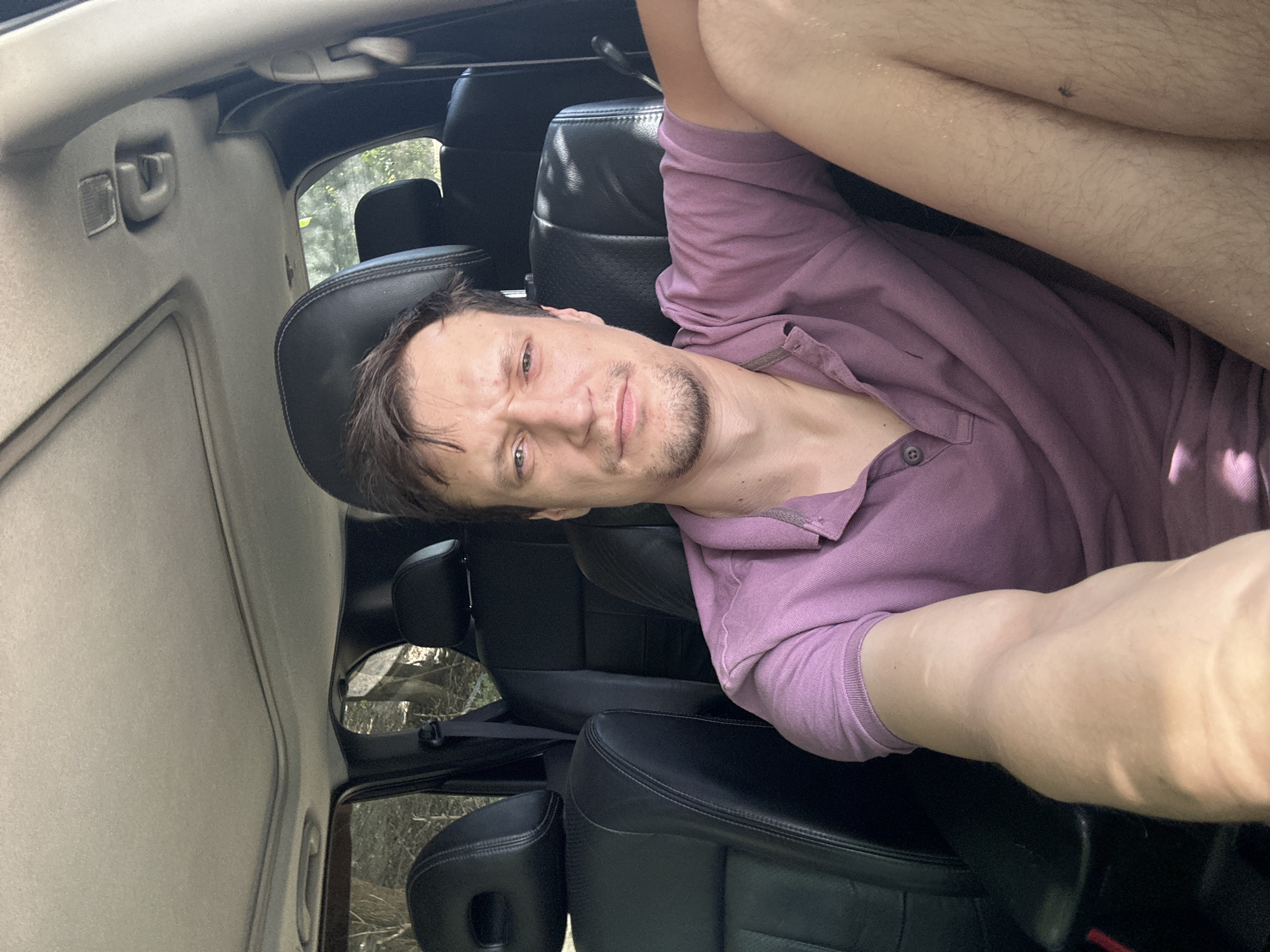 A man with short brown hair and a light beard is sitting in the driver's seat of a car, taking a selfie. He is wearing a purple polo shirt. The car's interior is visible, including the black leather seats, the sunroof, and the dashboard. Sunlight streams in from the side, casting shadows on the man's arm and chest. The background outside the car appears to be a wooded area.