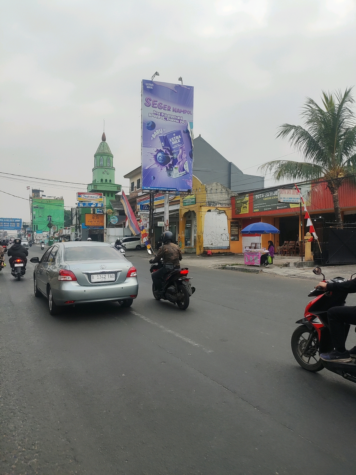 The image captures a street scene in Bekasi, Indonesia, on an overcast day. In the foreground, a silver Toyota Vios sedan with license plate B 1362 TAB is driving on the right side of the road. To the right of the sedan, a motorcyclist on a black scooter is also traveling in the same direction. Further ahead, other vehicles, including motorcycles and cars, are visible on the road.
The urban environment features various buildings lining the street. A prominent green mosque with a distinctive dome and minaret stands out on the left side of the image. To the right of the mosque, a large billboard advertising "ULTRA MILK" is displayed. Next to the billboard, a building with a red facade has a sign that reads "PUTRA JATI JEPARA FURNITURE & DESAIN". A small food stall with a blue umbrella is set up in front of this building, with a woman sitting under it. Palm trees are also present in the scene, indicating a tropical climate. The sky is cloudy, and the overall lighting suggests diffused daylight. The road appears to be paved with asphalt and shows some signs of wear and tear, including cracks.
