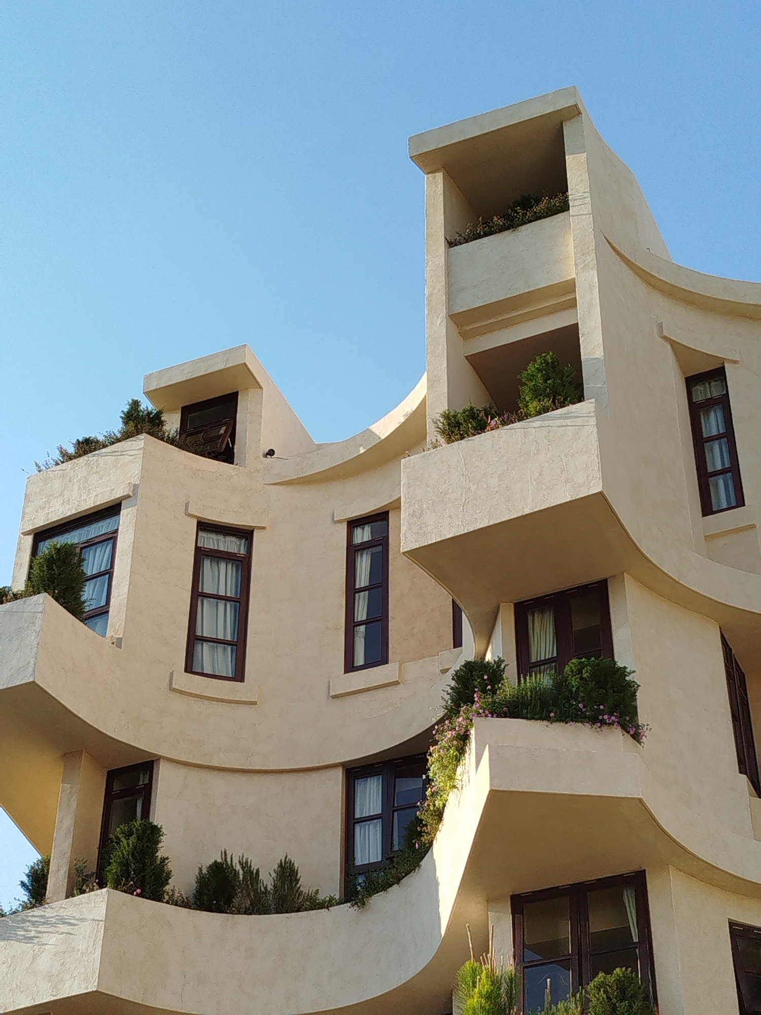 The image shows a multi-story building with an unusual architectural style. The exterior of the building is a light beige color, and it features curved walls, multiple balconies, and dark-framed windows. There are various plants and greenery on the balconies and rooftop areas. The sky is a clear, bright blue. The location appears to be Da Lat, Vietnam, based on the architectural style and vegetation, though this cannot be definitively confirmed from the image alone.