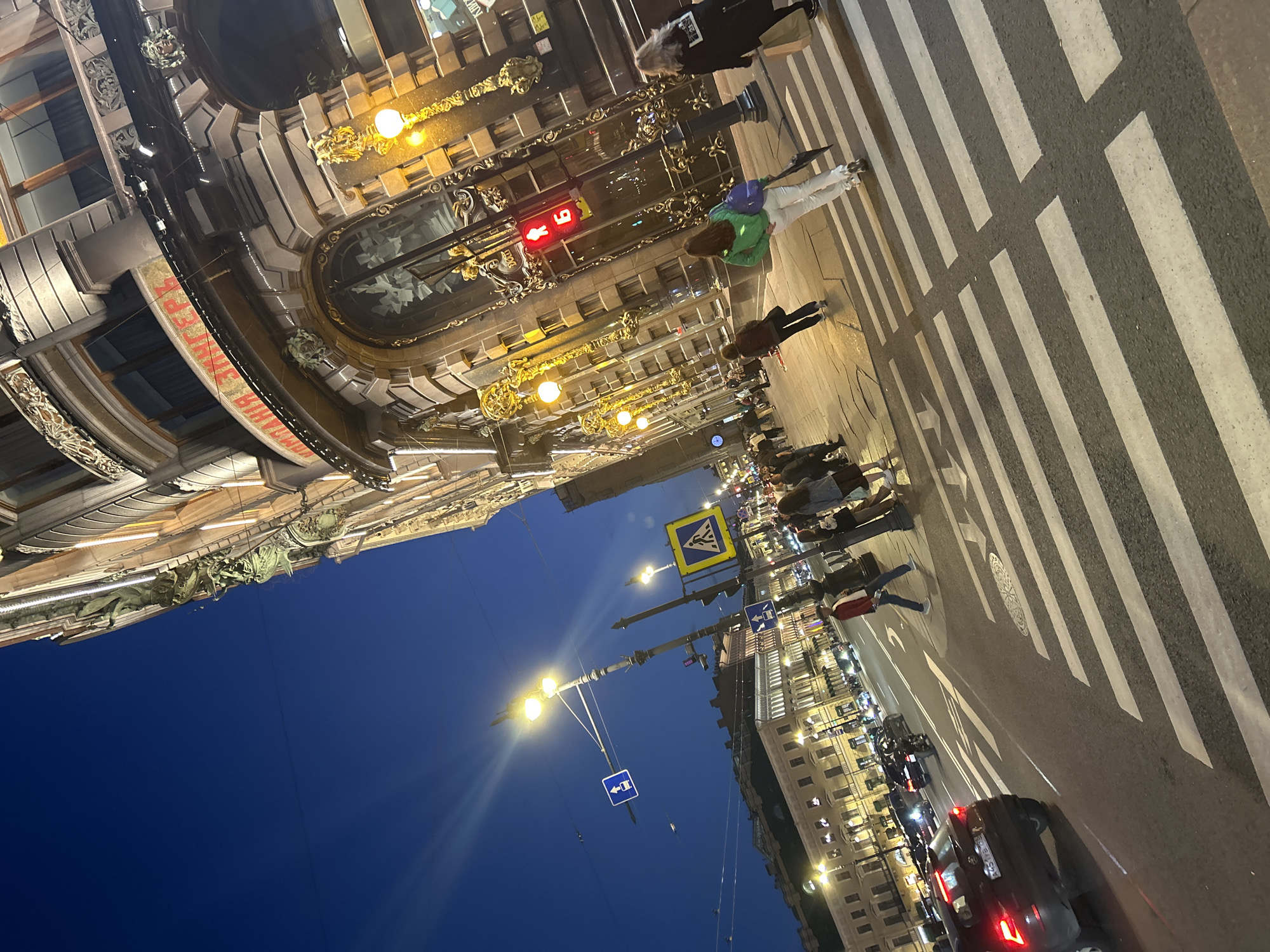 An eye-level, medium shot captures a street scene at dusk in an urban environment. The foreground is dominated by a pedestrian crosswalk with prominent white stripes against dark asphalt. Several people are visible crossing the street or walking on the sidewalk. To the right, a traffic light shows a red pedestrian signal. The background features a grand, ornate building with multiple stories, illuminated by warm, yellow lights. The building's facade is decorated with intricate architectural details and sculptures. Across the street, another row of buildings with illuminated windows lines the street. Several cars and a motorcycle are present on the road, and streetlights illuminate the scene. The sky above is a deep, dark blue, indicating the transition from day to night.