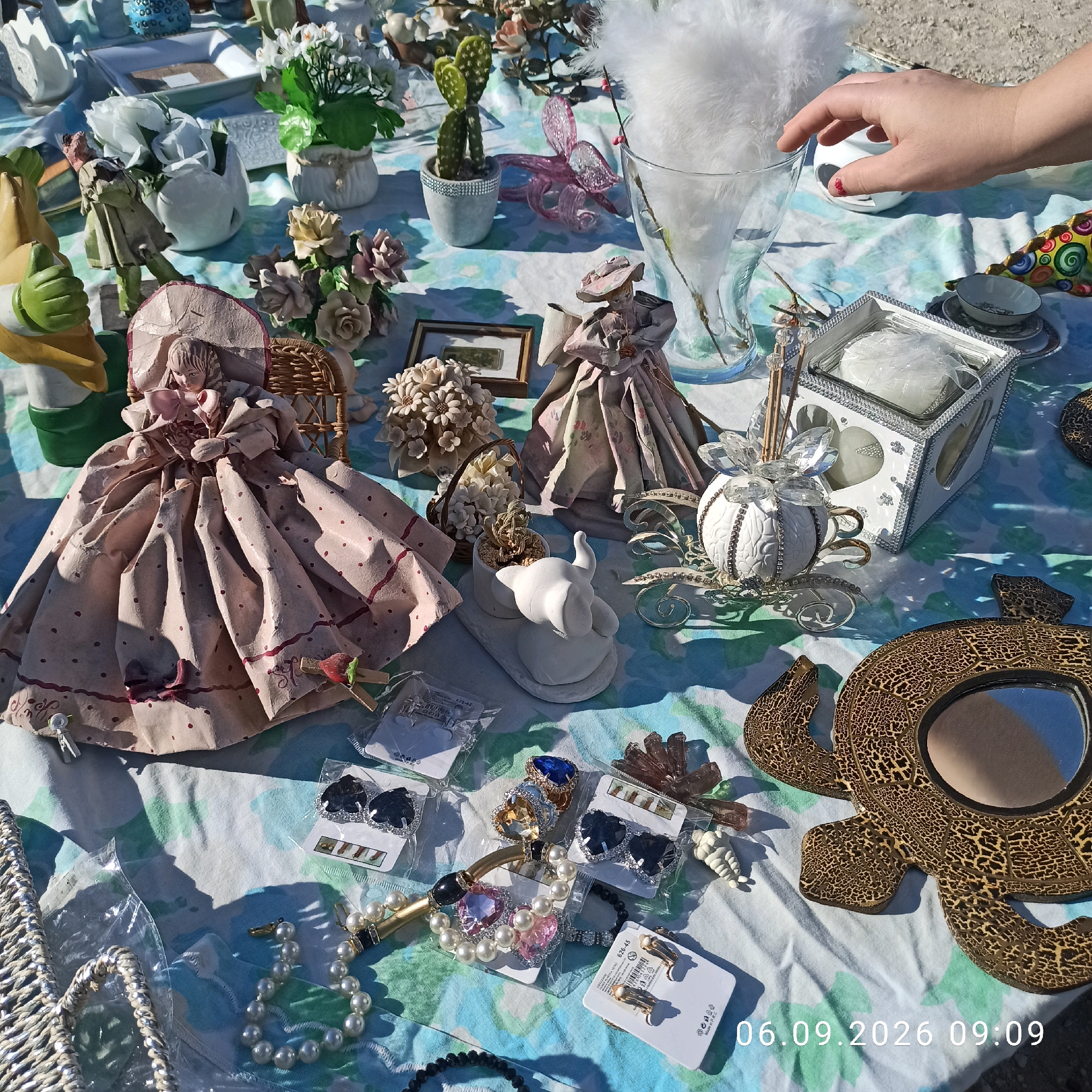 The image displays a close-up, high-angle shot of a table laid out with various decorative items, likely at an outdoor market or flea market. The surface is covered with a light-colored cloth featuring a floral pattern in shades of blue, green, and white. The background consists of a sandy or gravelly surface, suggesting an outdoor setting. The objects include a large, elaborately dressed doll in a pink, polka-dotted gown seated in a wicker chair, a white ceramic elephant figurine on a base, a cactus in a small pot, and several pieces of costume jewelry, including pearl necklaces and earrings. Other items visible are a glass vase filled with white feathers, a decorative white box with crystal embellishments, a small framed picture, and a gold-colored mirror with a cracked pattern resembling a turtle. In the upper right corner, a person's hand is visible, reaching towards the glass vase. A timestamp "06.09.2026 09:09" is superimposed on the lower right portion of the image. The scene appears to be set in Sannicandro di Bari, Italy, given the user's prompt.