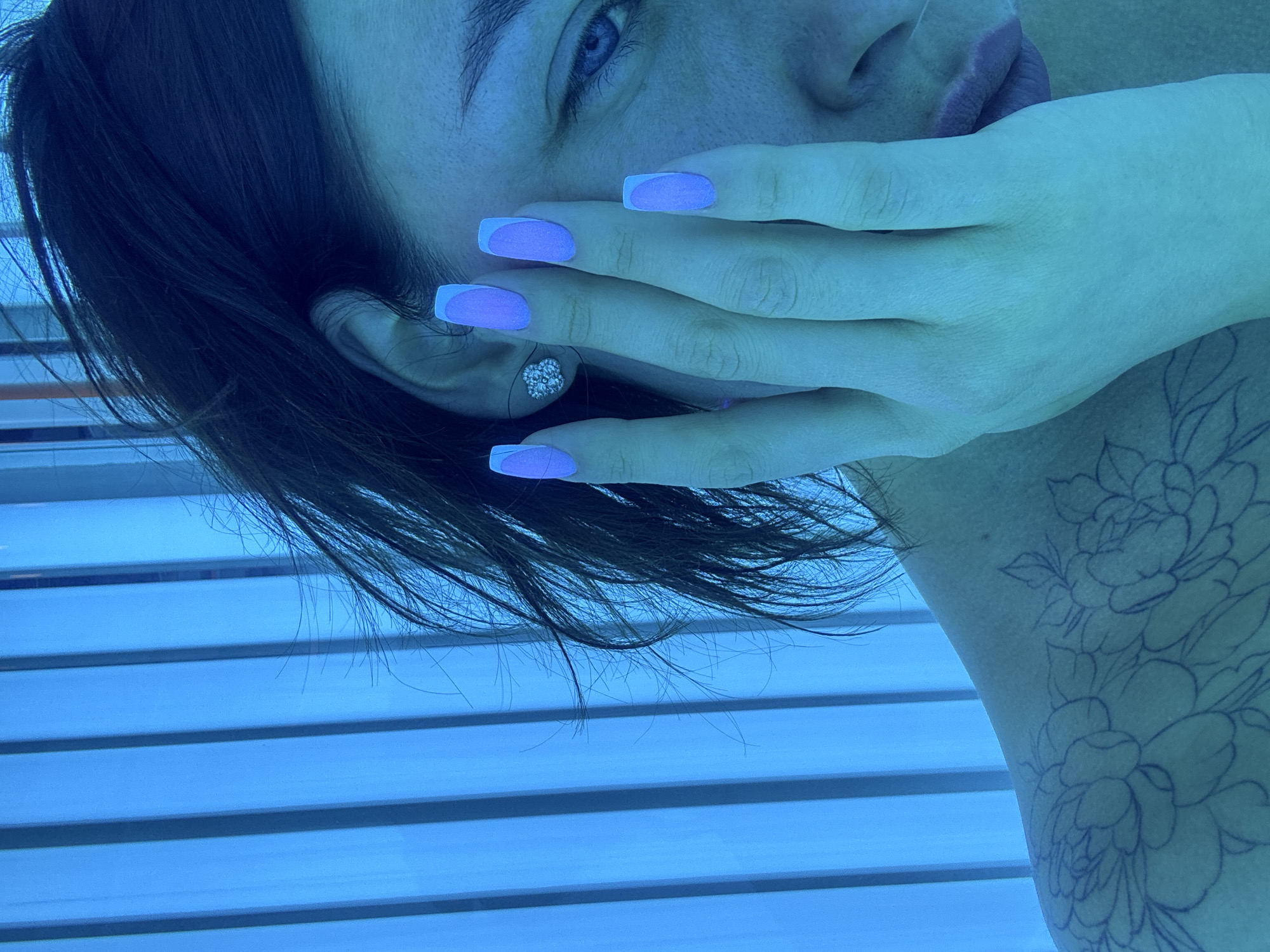 A woman with light skin, blue eyes, and dark hair is depicted in the image. She is lying in what appears to be a tanning bed, judging by the horizontal blue lines in the background. Her left hand is partially covering her face, displaying long fingernails painted with a lavender color and white tips. A small, possibly diamond, earring is visible in her left ear. There's a floral tattoo on her upper arm or shoulder. There's no information to indicate a location of Chaikovsky, Russia.