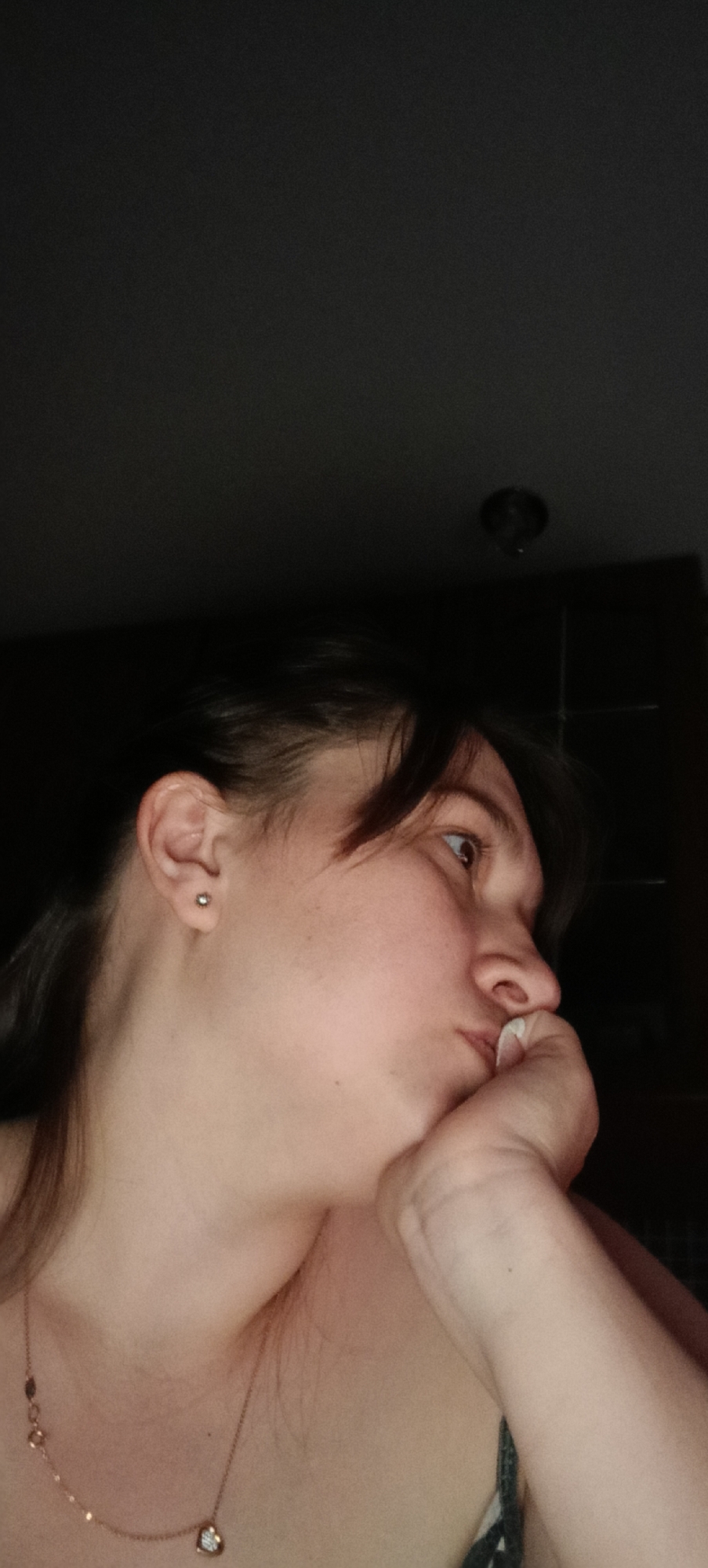 A close-up, profile shot shows the left side of a person's face, with their hand resting on their chin and mouth. Their eyes are looking off to the right, suggesting a pensive or thoughtful mood. The background is dark and nondescript, providing little context about the environment. There are no visible architectural elements or natural landscapes that would indicate a specific location within the city of Хохлы, Russia, or anywhere else. The lighting is dim, with the focus on the person's face and upper body. The person is wearing a necklace.