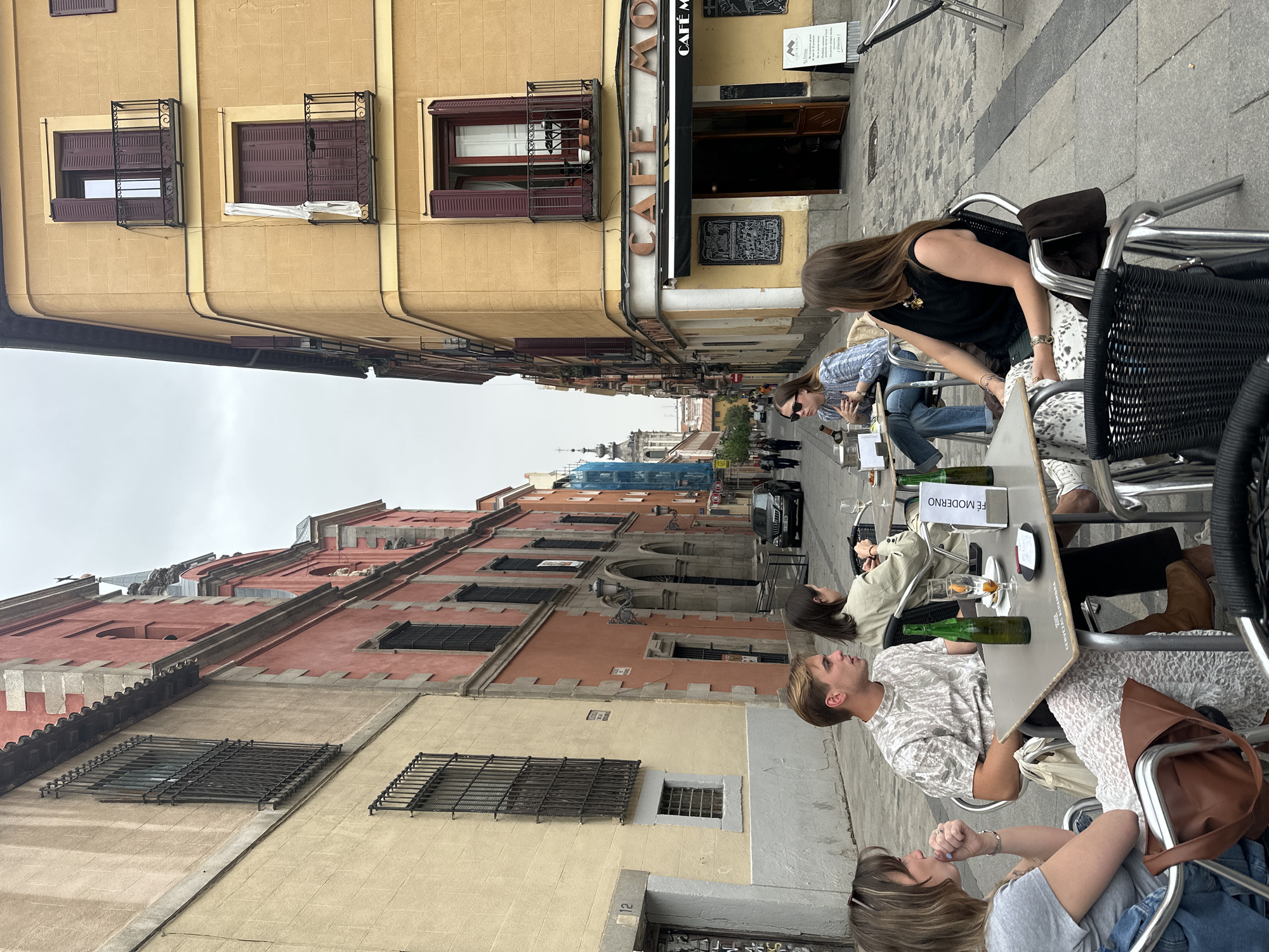 This image shows a street scene in Madrid, Spain. The buildings on either side of the street are old and have traditional Spanish architecture. On the right side of the street, a cafe with the name "CAFÉ MODERNO" on the sign is visible. Several people are sitting at tables outside, likely customers of the cafe. They are engaged in conversation and enjoying drinks. The street is narrow, and in the distance, a building with a spire covered with blue construction tarps is visible. The weather appears overcast.