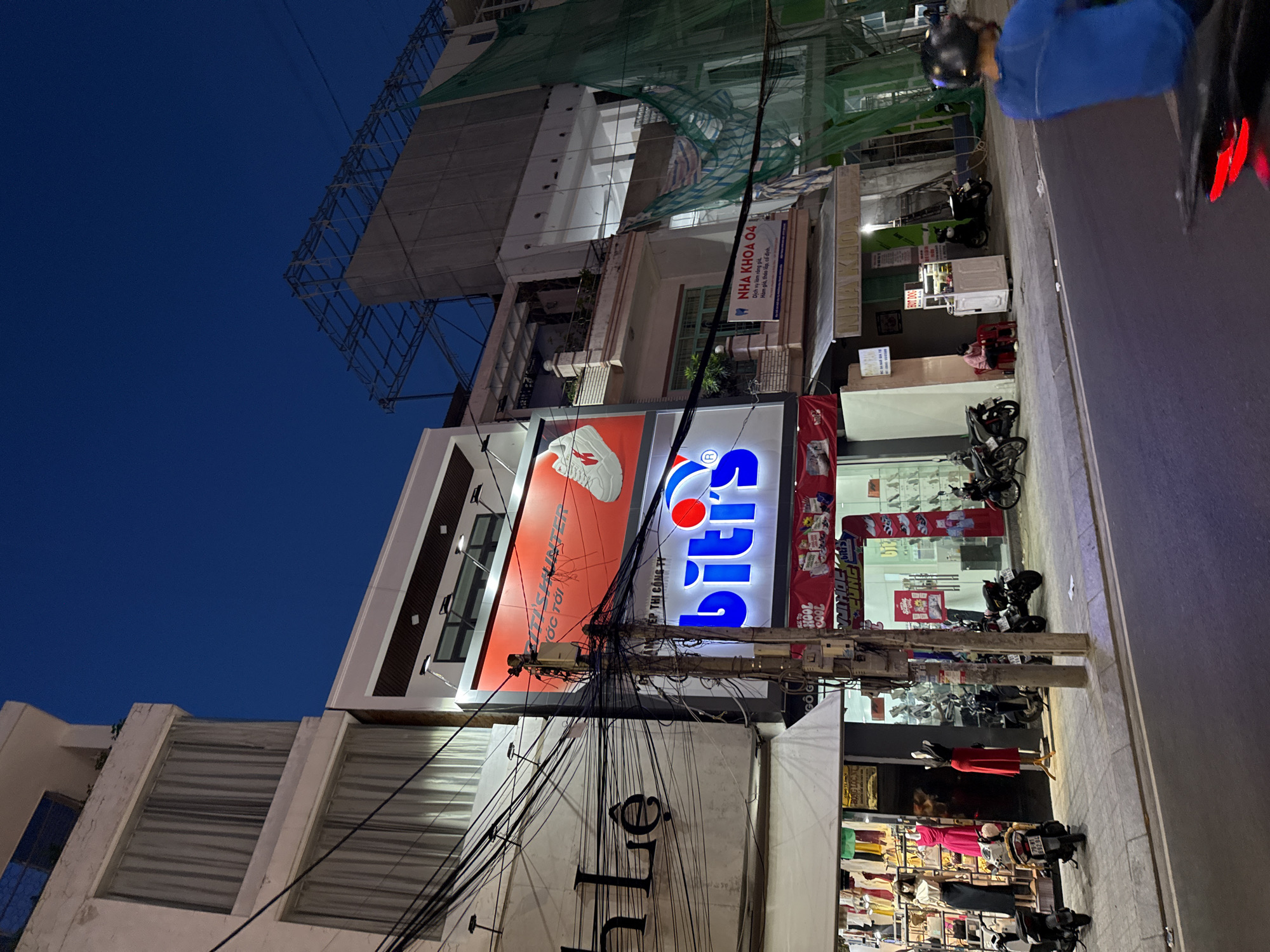 This is a photograph taken at night on a city street in Nha Trang, Vietnam. The scene depicts a street with several buildings lining the sidewalk, illuminated by artificial lighting. A motorcyclist, wearing a blue shirt and a helmet, is visible in the foreground, riding on the street.
The buildings on the left side of the street include a white building with signs that read "TH LE" in large black lettering. Next to it is a building with a large red advertisement for "FITNESS HUNTER" which features a white athletic shoe. Below this is a prominent blue and white sign for "bitis". Further down the street, on the right, is a building with a sign that says "NHA KHOA 04" (Dental Clinic 04).
The sidewalk in front of the stores is lined with parked motorcycles. Several mannequins displaying clothing are visible in the windows of a store on the left. A small food cart selling "HOT DOG" is situated on the sidewalk. A person is seated on a small red stool near the food cart.
The street is paved and appears to be a busy urban area. The sky above is dark, indicating nighttime. The overall impression is of a commercial street scene in a Vietnamese city after dark.