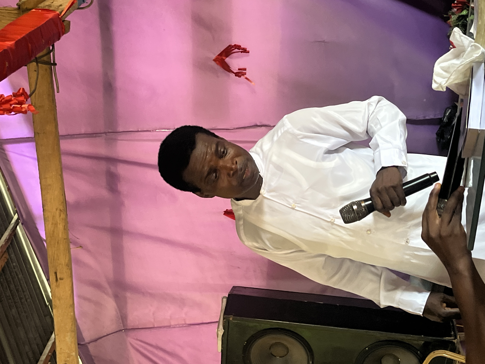 The image shows a man speaking or preparing to speak, rotated counter-clockwise. He is an adult male with dark skin and short, dark hair, wearing a white long-sleeved button-up shirt. He holds a black microphone in his left hand and looks slightly upwards and to his left with a neutral, focused expression.
To the man's right, another person's dark hand is visible, holding what appears to be a smartphone or small tablet. Below the man, a large, dark speaker unit with two visible speaker cones is placed. The background is composed of draped light purple fabric, likely serving as a temporary wall or backdrop for an event.
On the left side of the frame, there is a wooden pole with red, wrapped objects (possibly firecrackers or decorations) and red streamers or ribbons attached. Another small, red, claw-like decoration is affixed to the purple fabric wall. White fabric or paper is visible on a surface to the right, behind the man.
The scene suggests an indoor event, such as a religious gathering, public address, or performance, held in a temporary or simply adorned venue. The lighting indicates an indoor setting, and no specific time of day is discernible. There is no visible legible text. The context points to a community event in Iranla, Nigeria.