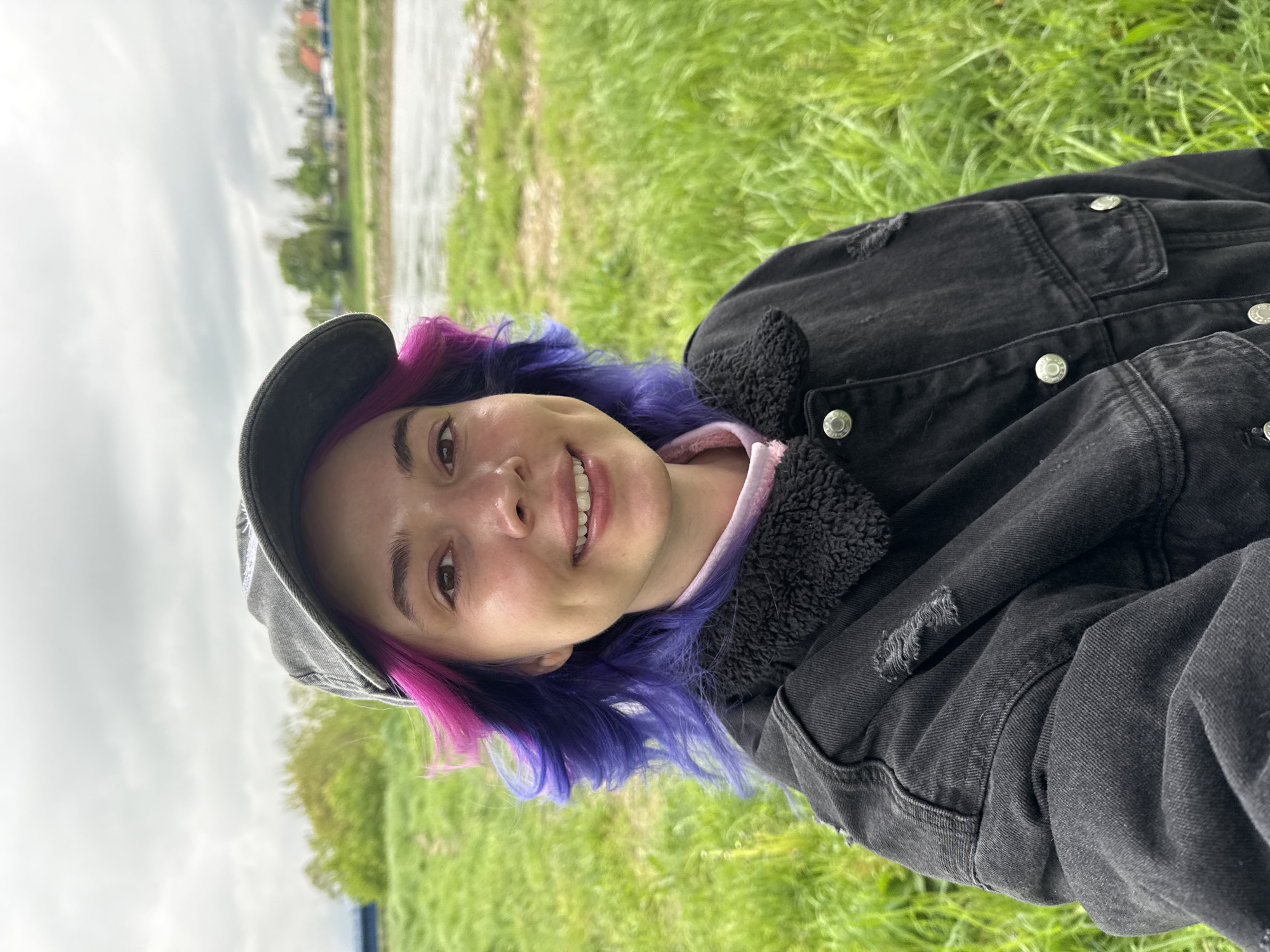 A young woman is lying in a grassy area on a cloudy day. She is wearing a gray cap and a black denim jacket with a fleece collar. Her hair is dyed in purple and pink shades. She is smiling. In the background, there is a body of water and some buildings. The image was taken outdoors in the City of Zagreb, Croatia.