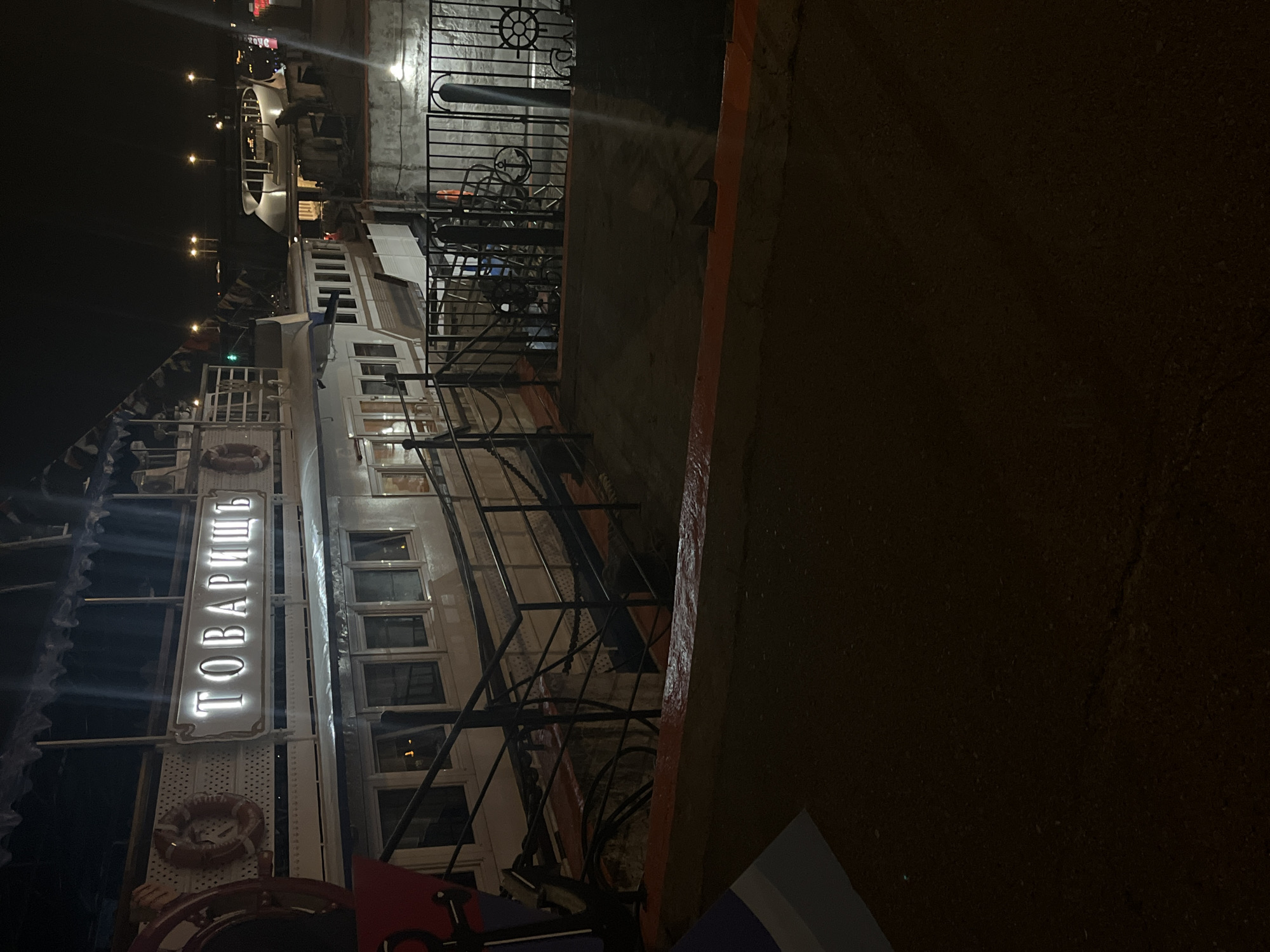 The image shows a multi-story boat named "Tovarish" docked at a pier at night in an urban environment. The boat is illuminated with lights that outline the Cyrillic name "ТОВАРИЩ" on its side. Multiple windows are visible along the boat's exterior. A black iron fence runs along the pier's edge, separating the boat from a wall. The surrounding environment is dimly lit, suggesting a nighttime setting in Unknown, Unknown.
