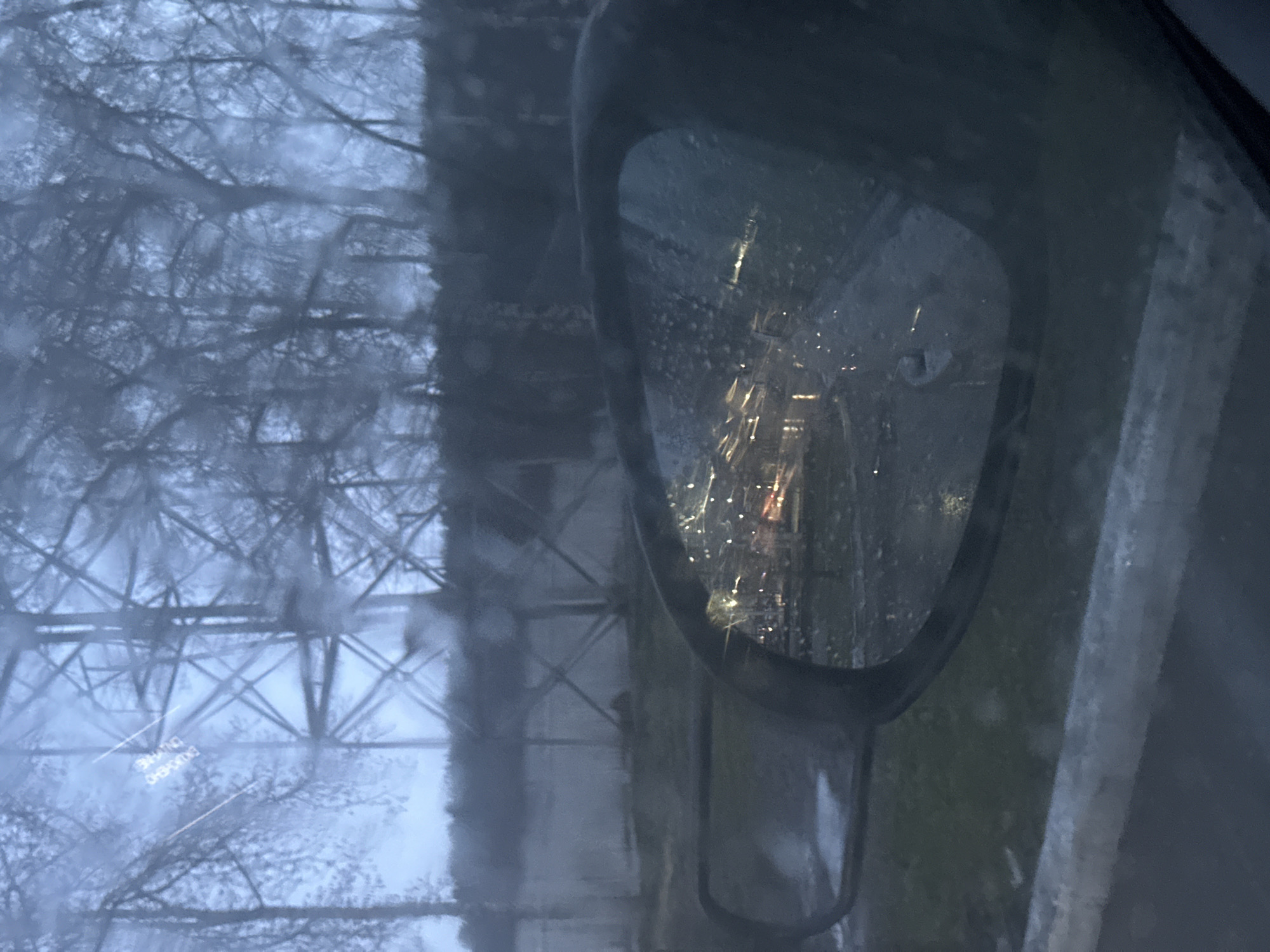 The image appears to be taken from inside a vehicle, possibly on a rainy or misty day. The visibility is reduced due to moisture on the glass. In the side view mirror, there is a blurred reflection of what appears to be a road at night with streetlights. Looking through the main window, a large metal transmission tower is visible, with bare branches of trees in the background. There also appears to be a sign, but the text is illegible. Since the location is Chaikovsky, Russia, the transmission tower is most likely part of the infrastructure related to energy distribution within the city. No people or actions are discernible. The overall environment seems urban.