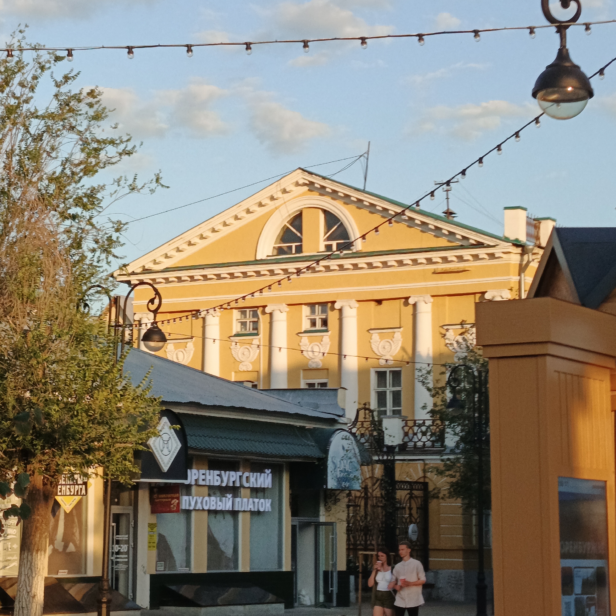 The image depicts a street scene in Orenburg, Russia, during what appears to be late afternoon or early evening, given the warm, golden light casting shadows. In the background, a large, ornate yellow building with classical architectural elements stands prominently. It features a semi-circular window at the apex, decorative cornices, and columns. In the middle ground, a smaller, dark-roofed building with a sign that reads "ОРЕНБУРГСКИЙ ПУХОВЫЙ ПЛАТОК" (Orenburg Downy Shawl) is visible, suggesting a retail establishment. Adjacent to this is a tree with dense green foliage.
In the foreground, a street lamp with a distinctive curved arm and a dark, globe-shaped fixture hangs from a string of lights that stretches across the scene. Another street lamp, similar in design, is visible on the left. The ground is paved, and a couple, a man and a woman, are walking in the middle ground, both holding cups. The man is wearing a light-colored shirt and dark trousers, while the woman is in a white top and shorts. They appear to be casually strolling. The overall environment suggests a historic urban setting, possibly a pedestrian street or shopping area. The sky above is blue with scattered, soft white clouds.