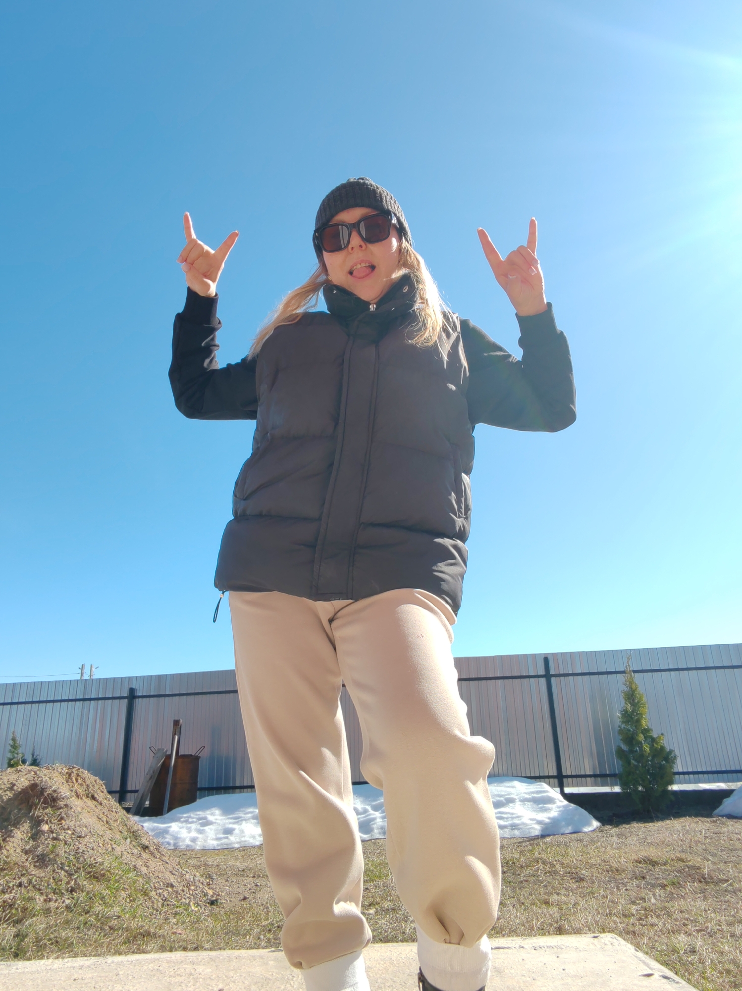 A woman stands outside on a sunny day in Chaikovsky, Russia, making a rock-and-roll hand gesture with both hands. She is wearing a black beanie, sunglasses, a black vest over a black long-sleeved shirt, and beige sweatpants with white socks. She is sticking her tongue out. In the background, there is a metal fence, a small evergreen tree, a pile of dirt, a few patches of melting snow, and a blue sky.