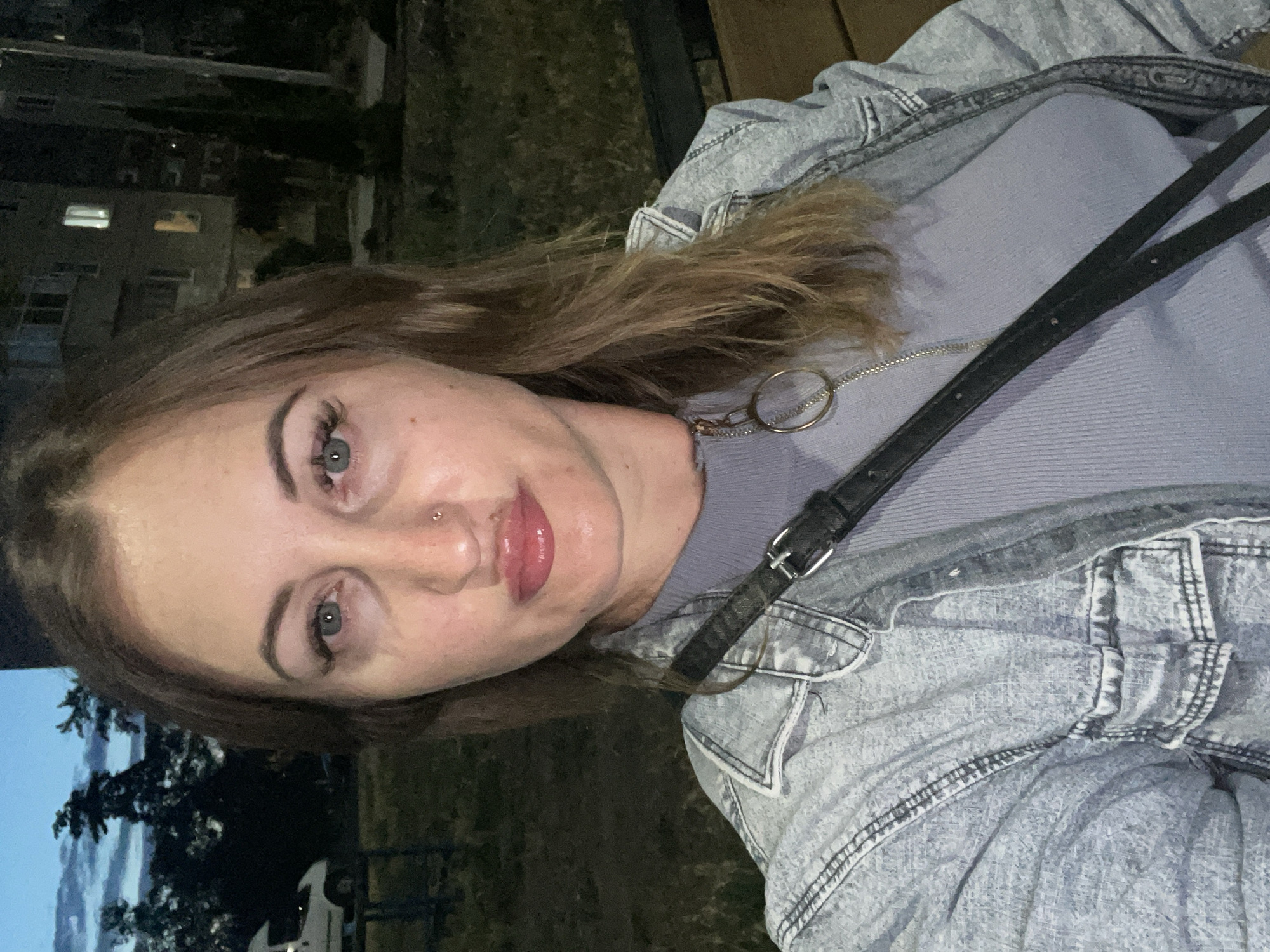 A woman with shoulder-length brown hair and light skin is photographed from the chest up, outdoors at dusk. She has a small nose ring and her eyes are wide open and looking towards the camera. She is wearing a light purple, ribbed, long-sleeved shirt with a zipper at the neck. Over the shirt, she has on a light blue denim jacket. A black strap from a purse or bag crosses diagonally across her chest, with a silver buckle visible on the strap. In the background, there are trees silhouetted against a twilight sky with clouds. To the right, a multi-story building with illuminated windows is visible, suggesting an urban or suburban environment, possibly within the city of Zarichne, Ukraine, given the context. The overall lighting suggests it is either early morning or late evening.
