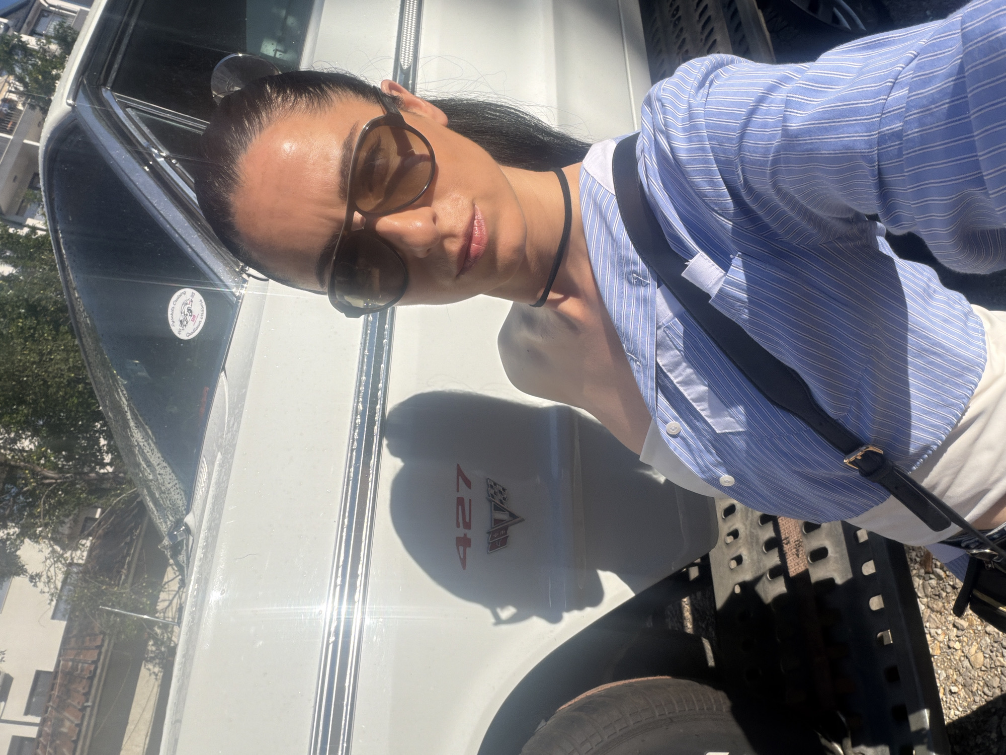 A woman wearing sunglasses, a blue and white striped shirt, and a black choker stands in front of a white car with a "427" emblem. The scene is outdoors, with sunlight casting shadows. The location is Pleven, Bulgaria. The car appears to be vintage.