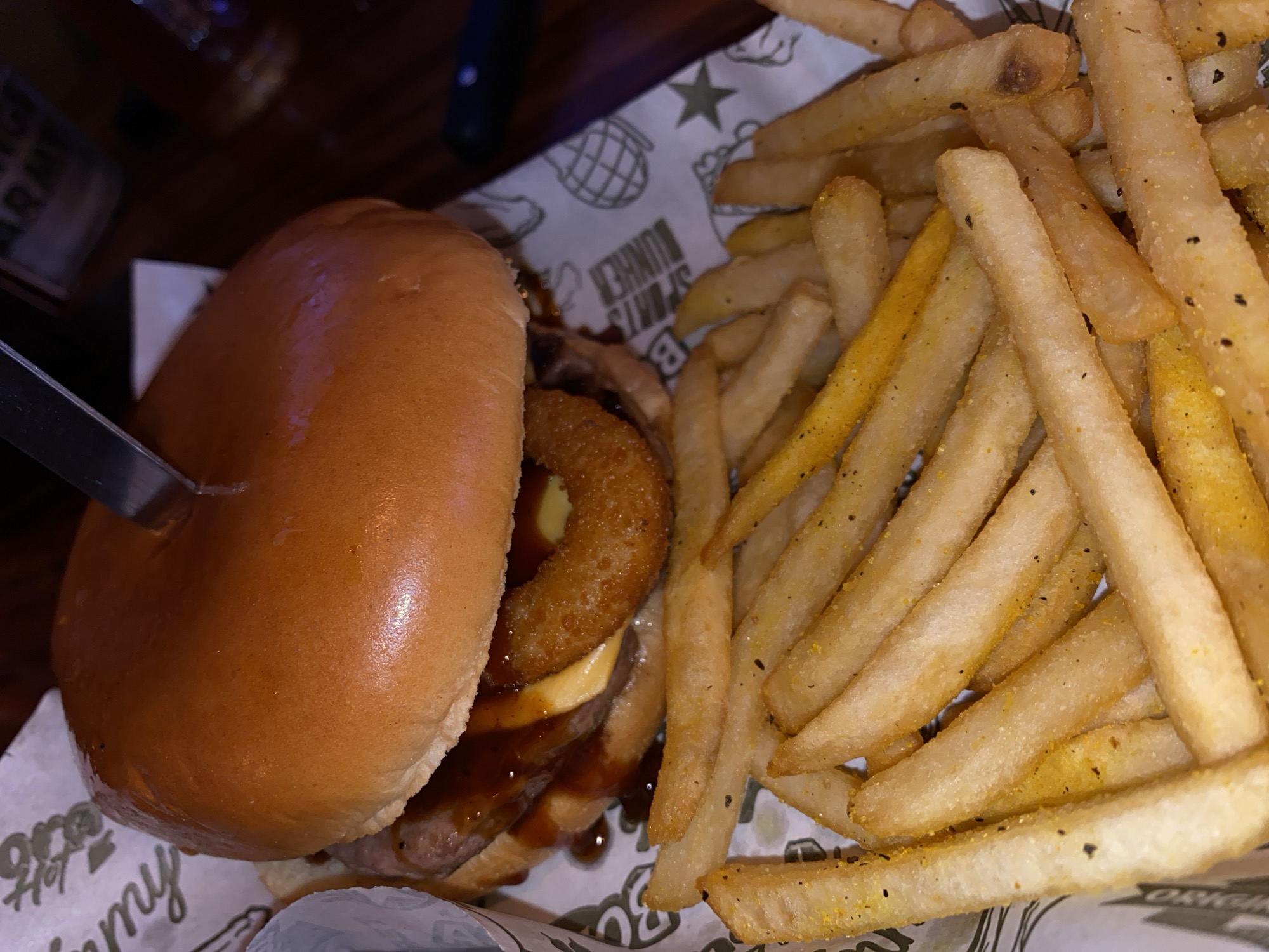 A high-angle shot depicts a hamburger and a pile of fries. The hamburger is topped with an onion ring and sauce, held together by a knife inserted into its left side. The fries are mostly light golden brown and seasoned with black and yellow seasonings. The food rests on a white and light-green printed paper liner inside a basket. The liner has a pattern of stars, grenades, and text including "SPORTS BURGER" and "Wings Army". The background appears to be a dark wooden table. The location and the city is unknown.