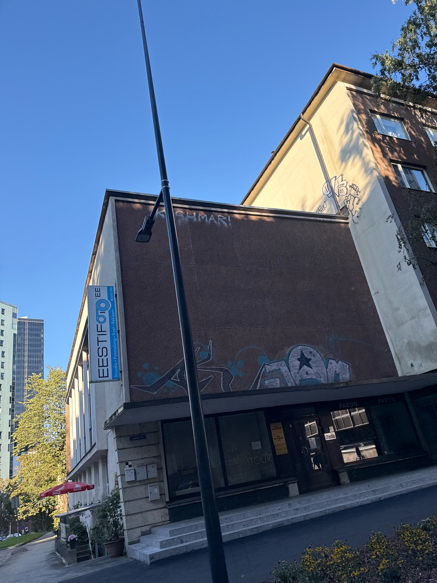 The image shows a multi-story building in Tallinn, Estonia, on a clear day. The building's facade features a mix of brown textured material and a lighter-colored section on the corner, with visible graffiti on both. A tall black lamppost stands prominently in the foreground, slightly to the right of the center. To the left of the building, a tall, modern, light-colored skyscraper is partially visible.
A vertical blue and white sign on the side of the building reads "EESTIFOTO" and lists services such as "Studio", "Fotosalong", "Kool", and "Reklaam", along with a phone number. Beneath this sign, the building has a ground-floor entrance with large windows. One window displays a sign that reads "STUDIO". Another sign on the glass indicates "COMATOUR AGENCY". Next to the building's entrance, there are several mailboxes mounted on a tiled wall, and a set of concrete stairs leads up to the entrance. An outdoor seating area with a red umbrella branded with "Coca-Cola" is visible to the left of the entrance, accompanied by potted plants. Further left, a tree with green foliage is prominent. In the background, another tall building with a darker facade is visible. The sky is a clear, bright blue.