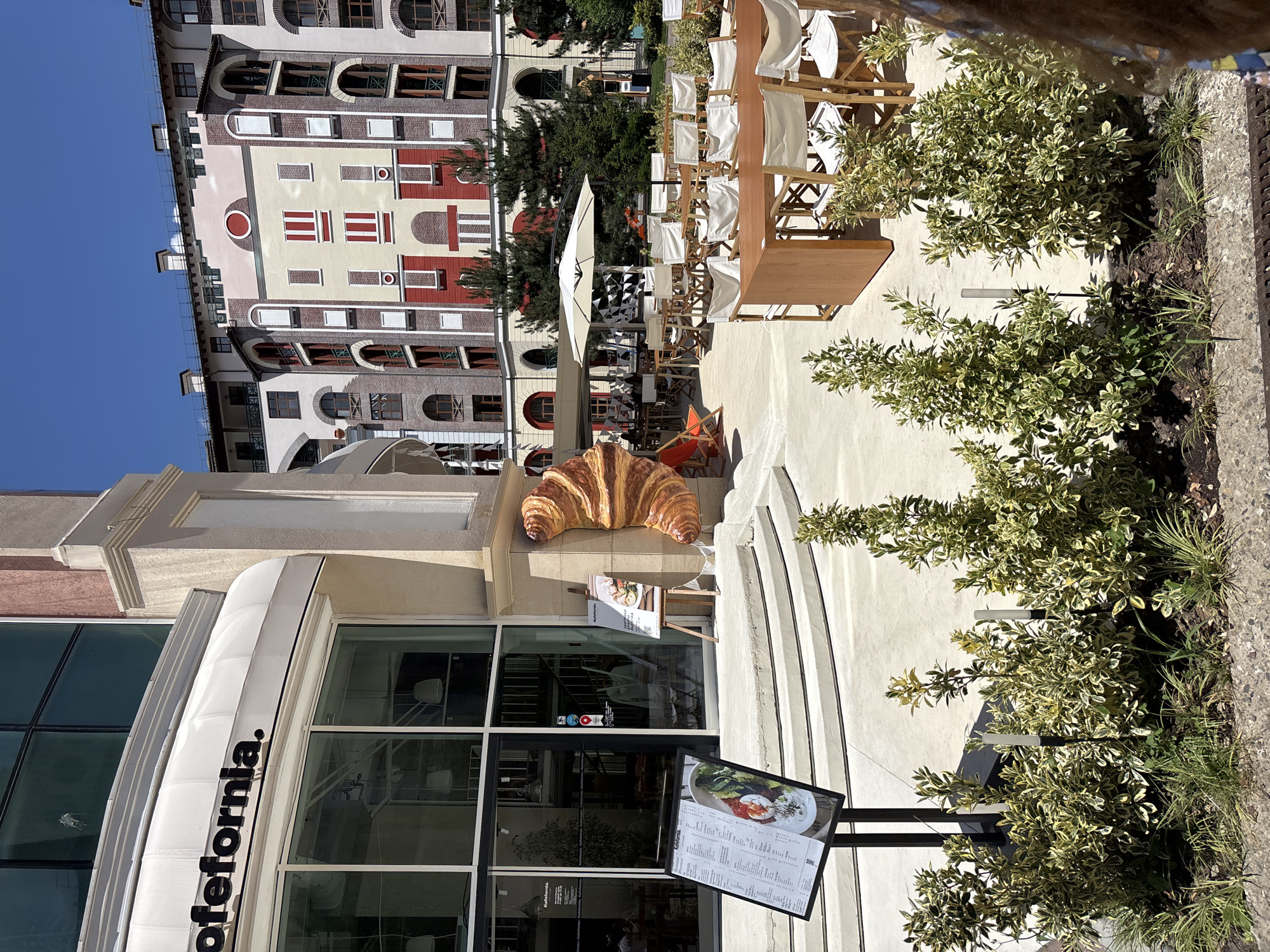 This image shows an outdoor seating area in front of a cafe named "Cafeformia" in Estosadok, Russia. The cafe has a modern facade with large glass windows and a white awning. A large, realistic sculpture of a croissant is prominently displayed next to the entrance. In front of the cafe, there are several steps leading up to the entrance, and a menu stand is placed on the left. The seating area features numerous wooden tables and white chairs, some of which are set up under a large white umbrella. In the background, a multi-story residential or hotel building with a colorful facade, featuring red, yellow, and white elements, is visible against a clear blue sky. Several evergreen trees are scattered around the seating area. The scene appears to be a sunny day, with shadows indicating bright sunlight.