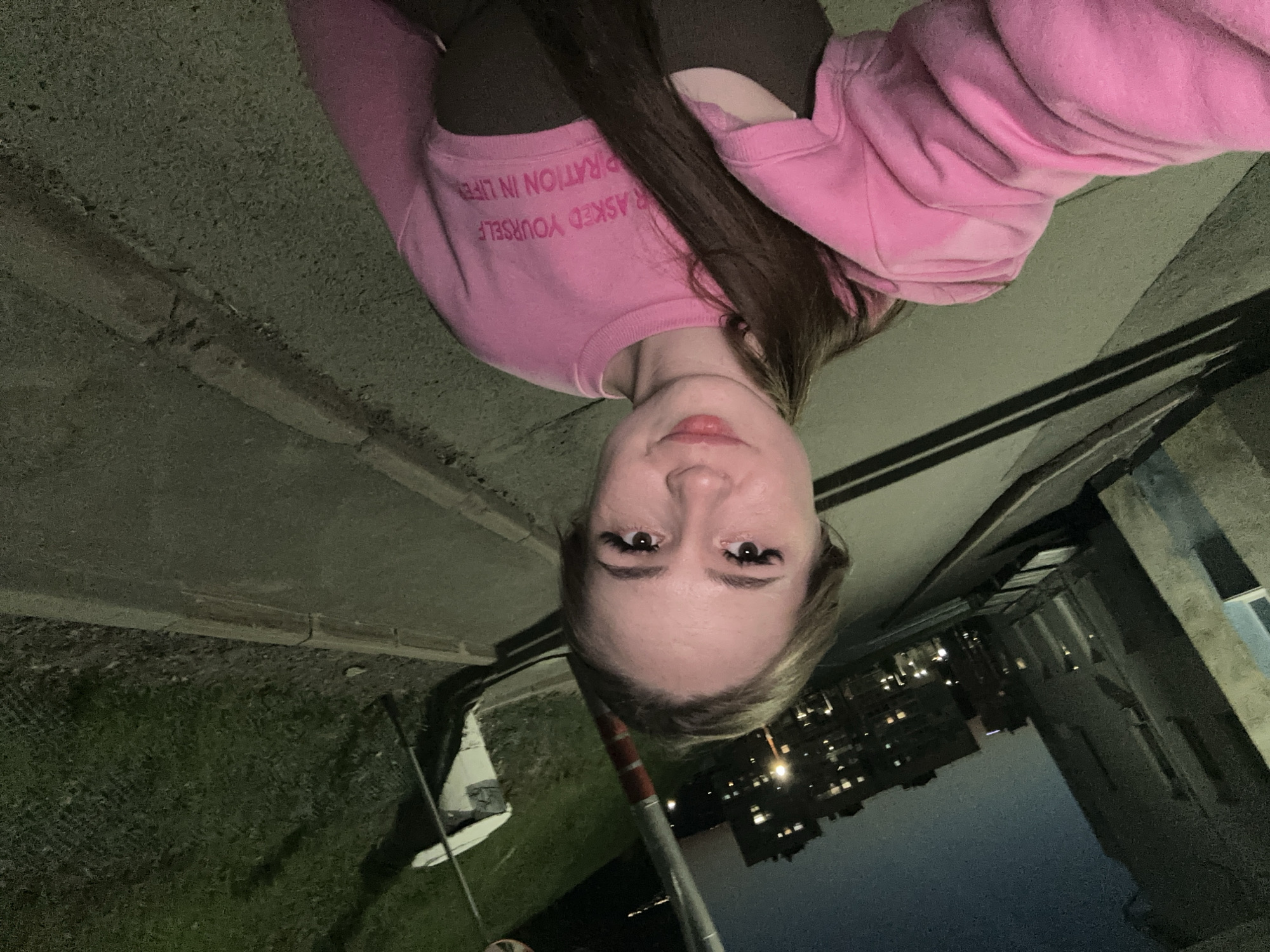 A young woman is depicted in an outdoor, nighttime setting, likely posing for a selfie. She has long, dark brown hair that falls over her shoulders and is wearing a bright pink crewneck sweatshirt over a dark top. Her facial features include dark eyes with prominent eyelashes and slightly parted lips with a hint of a smile. The text on her pink sweatshirt is partially visible, reading "ASKED YOURSELF RATION IN LIFE," suggesting part of a phrase like "HAVE YOU EVER ASKED YOURSELF WHAT IS YOUR ASPIRATION IN LIFE."
The background indicates an urban or suburban environment at night, with the dark sky contrasting with the illuminated windows of distant buildings. To the left of the woman, there is a grassy area adjacent to a concrete or asphalt path, with a white object, possibly a boat or a piece of equipment, and a fence visible within the grass. A red and white striped pole stands vertically in the mid-ground. The foreground consists of textured dark concrete or pavement. The overall lighting suggests artificial illumination from streetlights or nearby buildings. The image is captured from a slightly low angle, emphasizing the woman and her immediate surroundings.