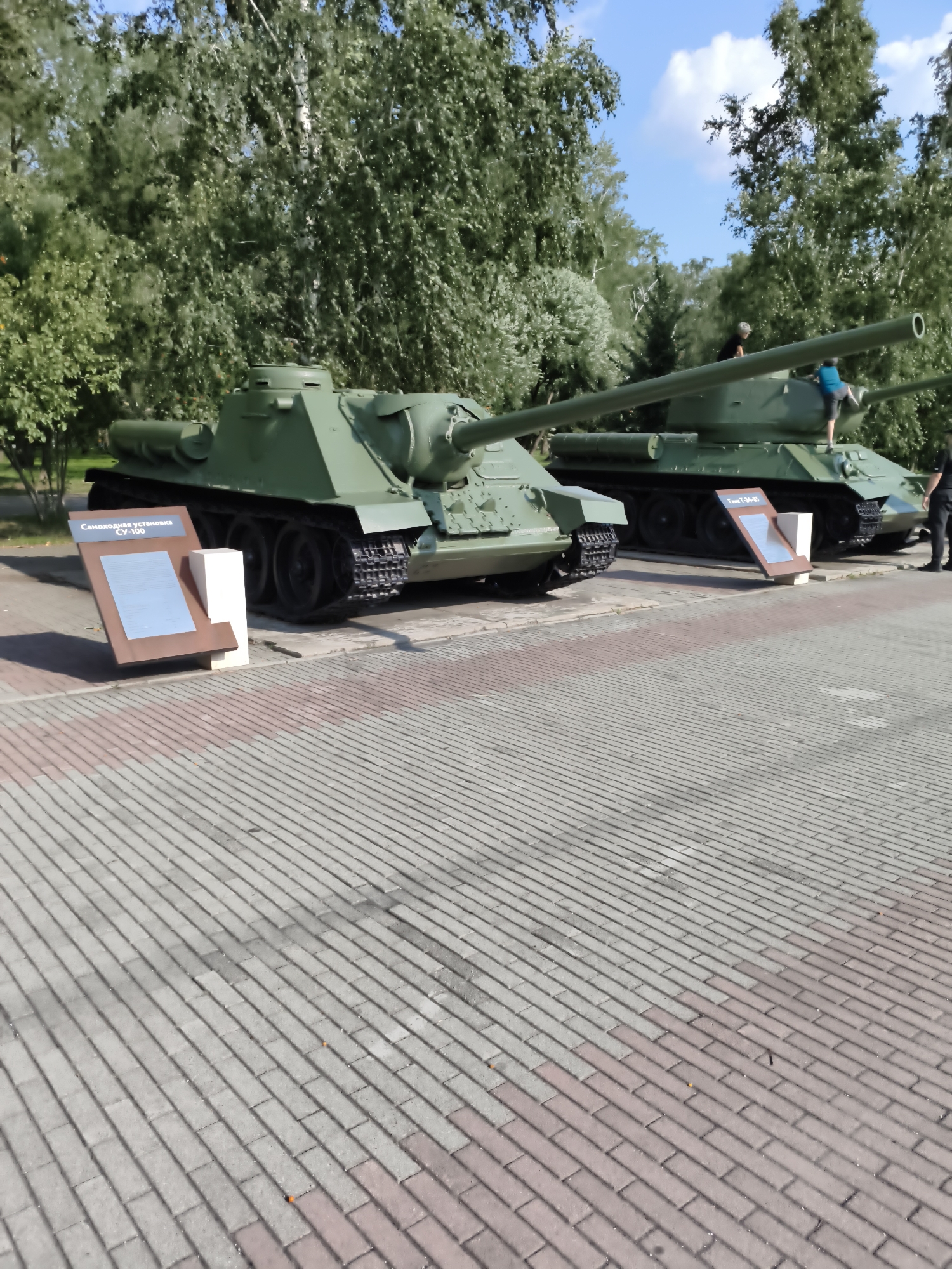 The image depicts two green military tanks, a SU-100 self-propelled gun in the foreground and a T-34-85 tank behind it, displayed on a paved area with trees and blue sky in the background. A person in a dark shirt is standing near the T-34-85. On the SU-100, a person in a blue shirt is climbing on the tank. A plaque is visible in front of the SU-100 with Russian text, and another plaque is visible in front of the T-34-85, also with Russian text. The scene is outdoors, likely in a park or museum dedicated to military history. The image was taken in Chelyabinsk, Russia, at the Victory Park.