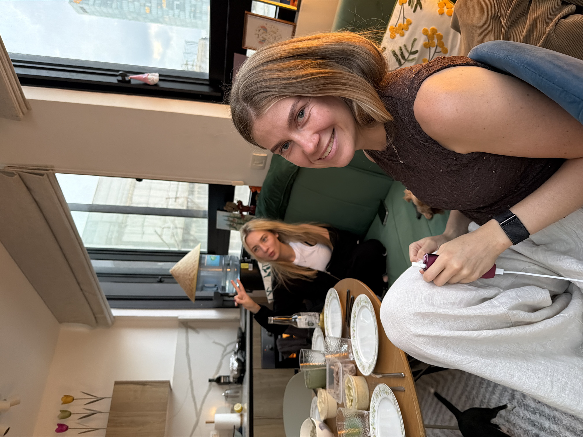 Two women are at a dining table with plates and glasses. One woman is seated and holding up two fingers. The other is holding a purple device. A partially visible black cat is under the table. A window shows a building outside. A beige Asian hat is hanging. The location is possibly Tel-Aviv, Israel.