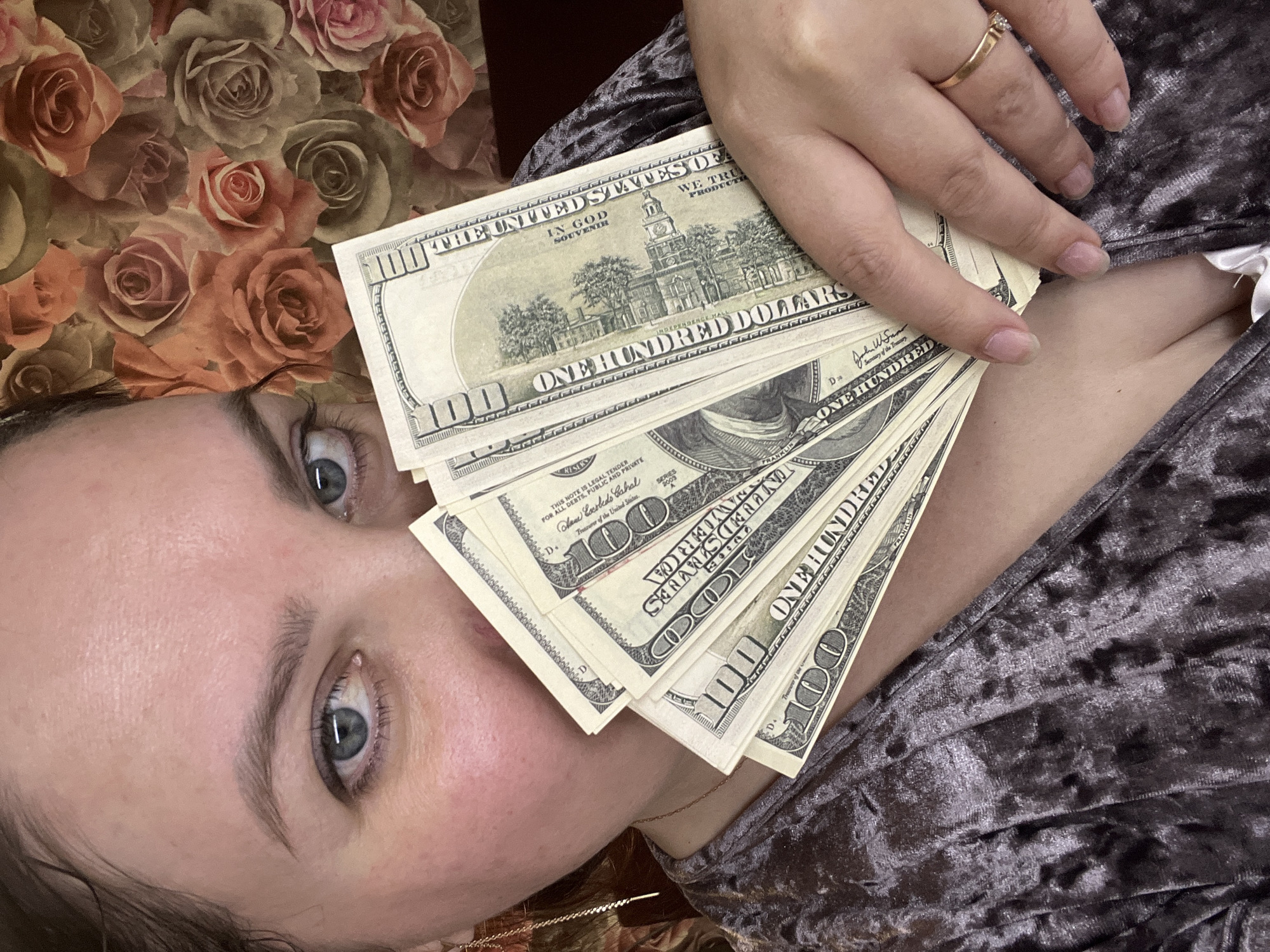 A woman lies on a surface with a rose pattern, wearing a textured garment, and holds a stack of $100 USD bills in her hand, partially covering her face. The woman is wearing a ring on her finger.