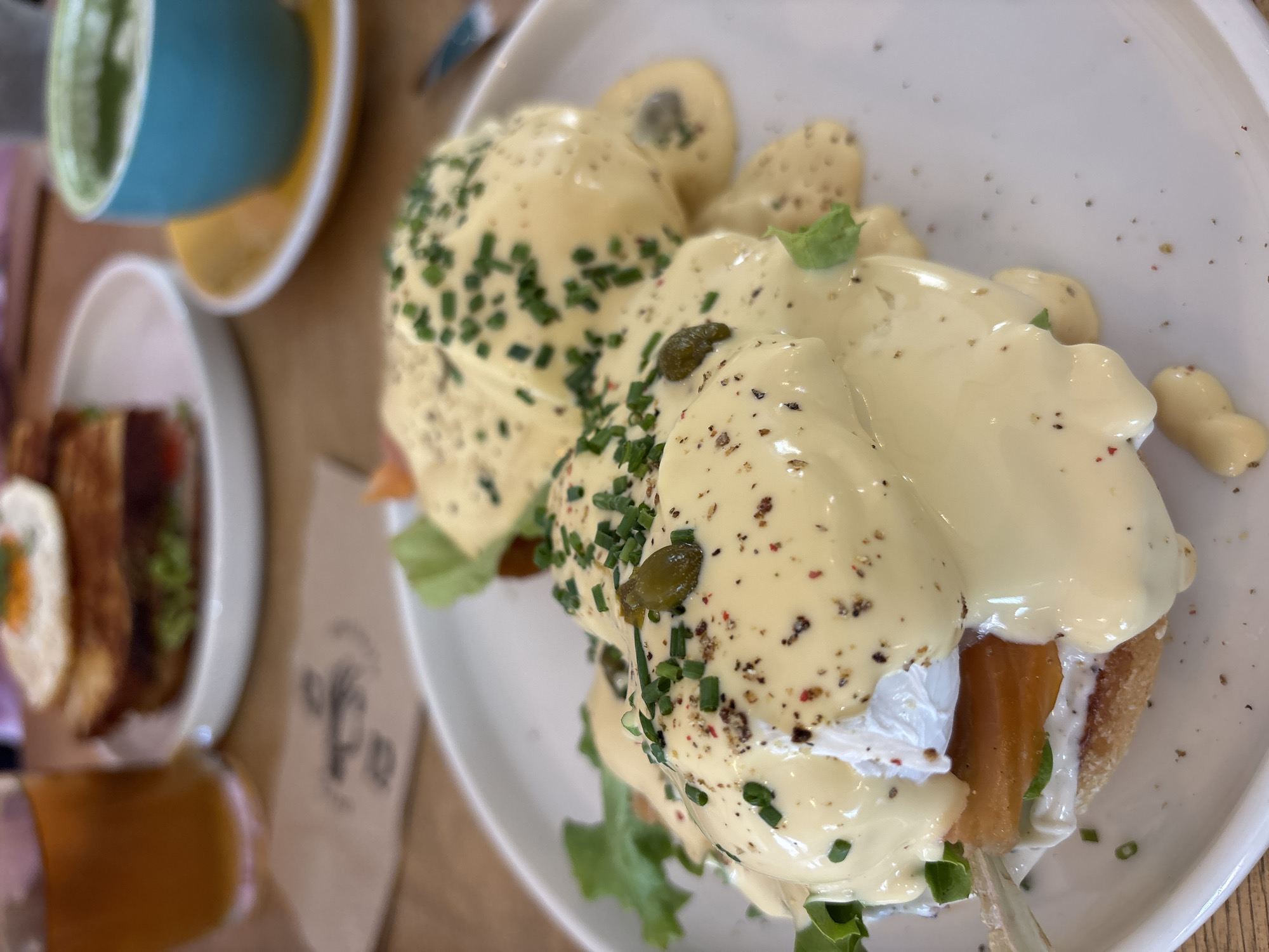 A close-up, slightly angled shot shows a plate of Eggs Benedict, topped with hollandaise sauce and herbs, sitting on a wooden table. Another plate with a different dish and a cup are partially visible in the blurred background. The location is indicated to be in the city of Varna, Bulgaria.